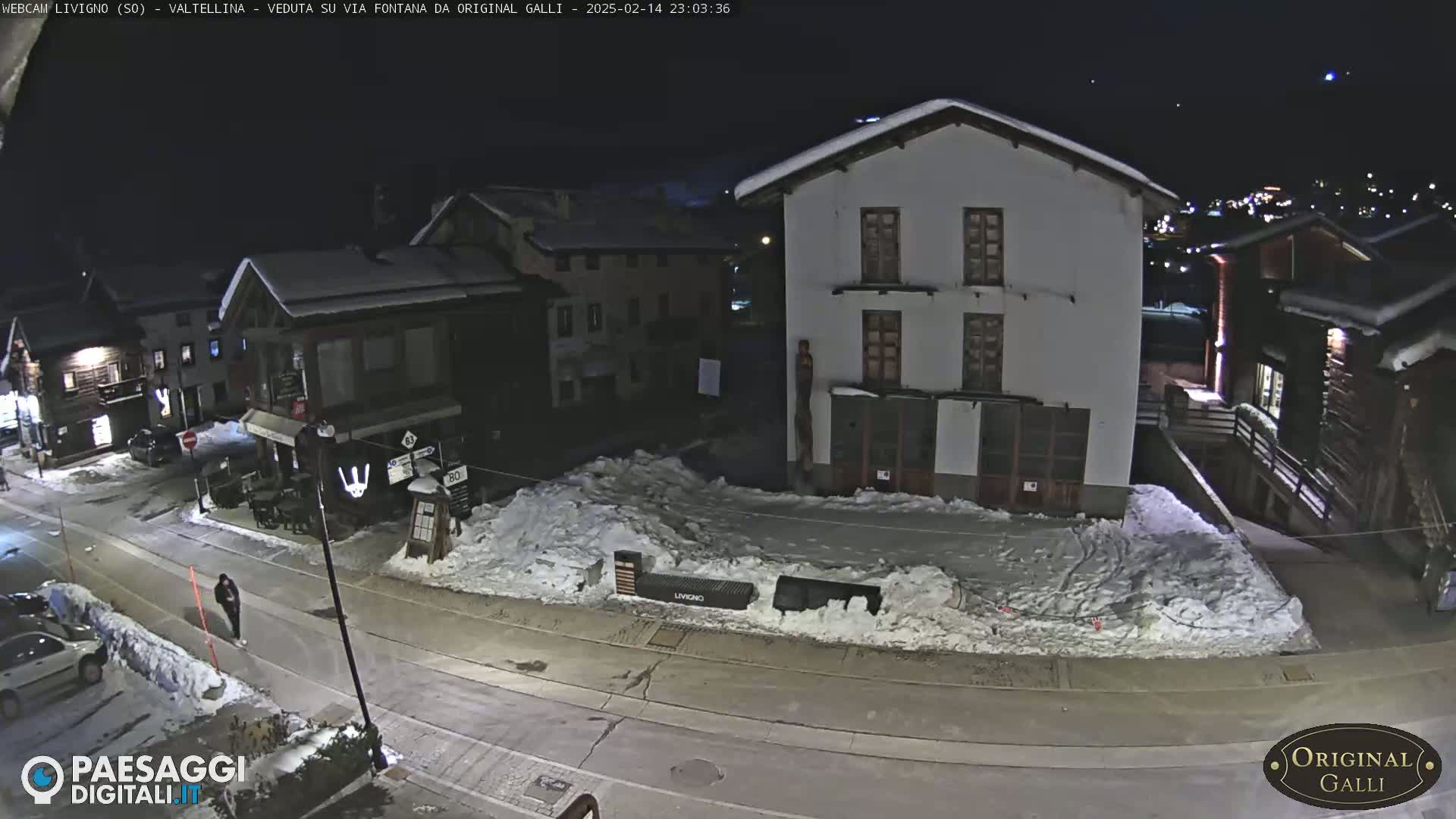 A snow-covered street scene at night, showing several buildings and a person walking.