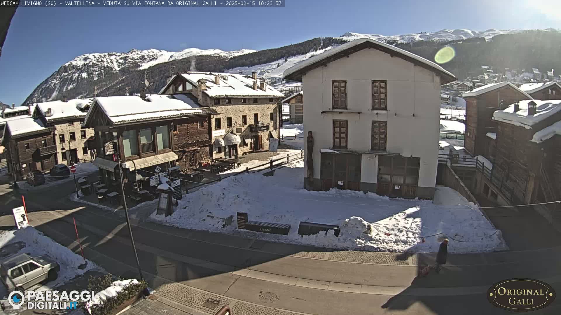 A sunny, snow-covered village square with several buildings, one of which is a large white structure, is viewed from above.