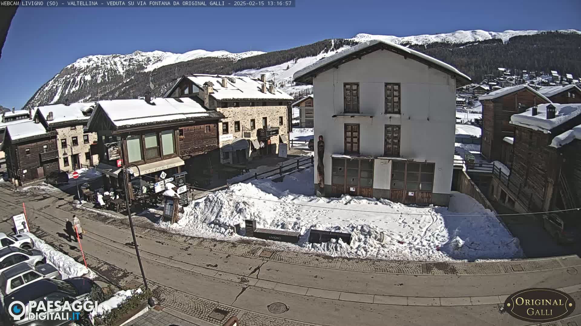 A snow-covered village square with several buildings, some cars parked along a street, and mountains in the background under a sunny sky.