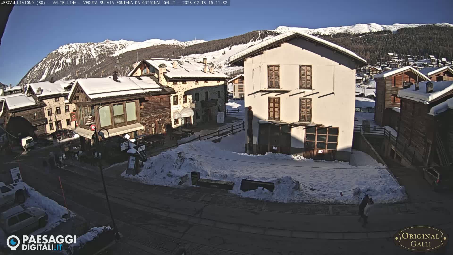 A snow-covered village square, nestled in a valley between snow-capped mountains under a clear blue sky, shows several buildings with snow on their roofs.