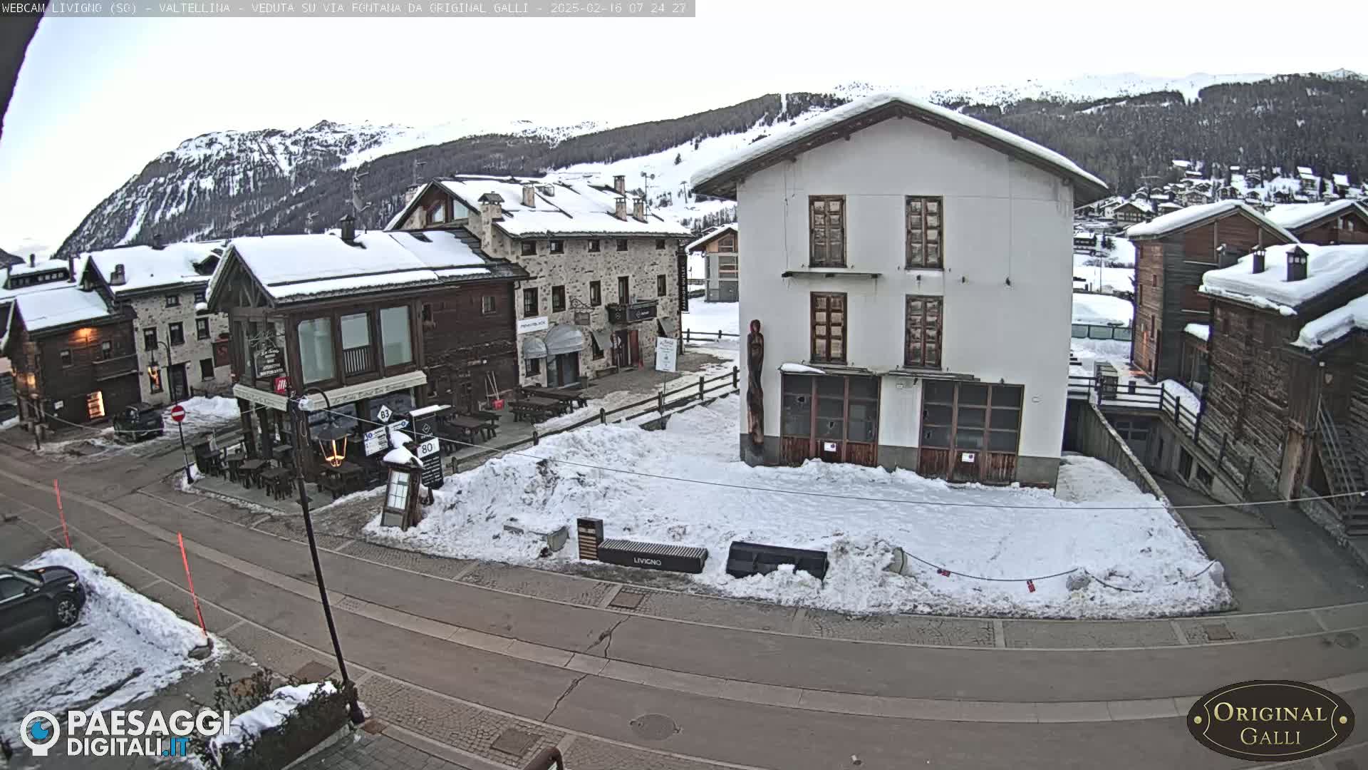 A snow-covered village square in a mountainous region, with several buildings and a street visible.