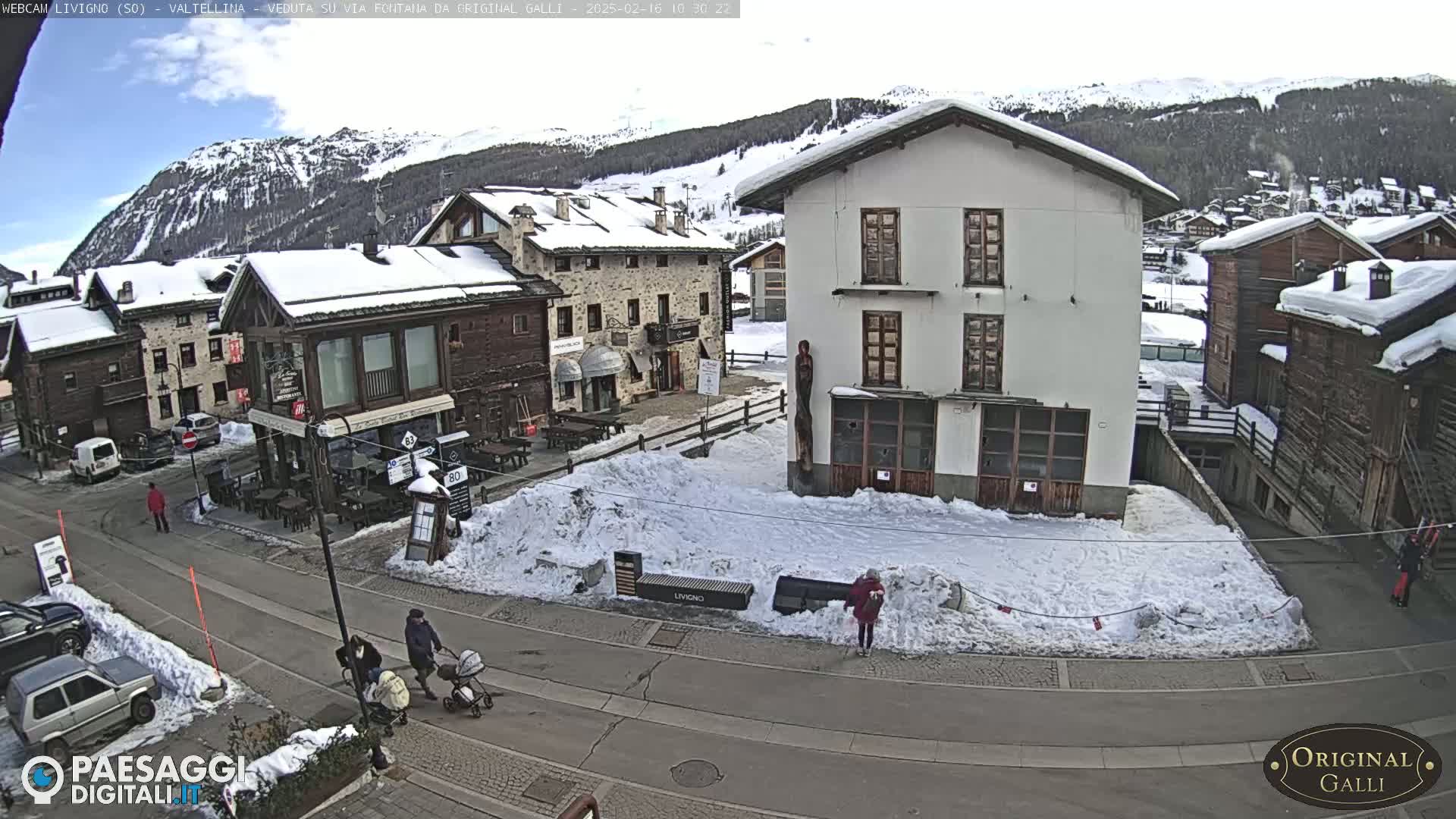 A snow-covered village street scene shows several buildings, some pedestrians, and parked cars under a partly cloudy sky.