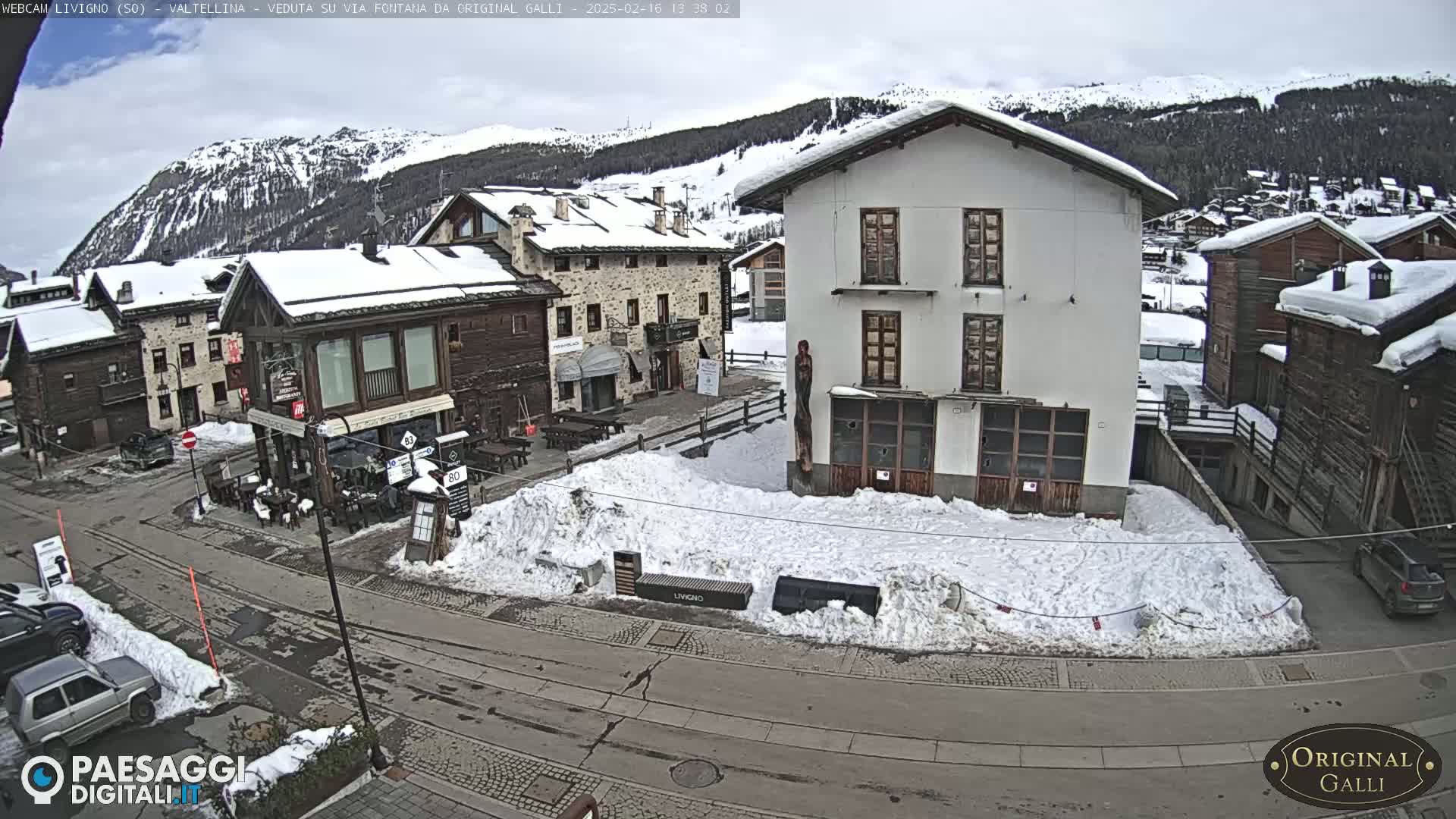 A snow-covered village square, surrounded by snow-dusted buildings and mountains under a cloudy sky.