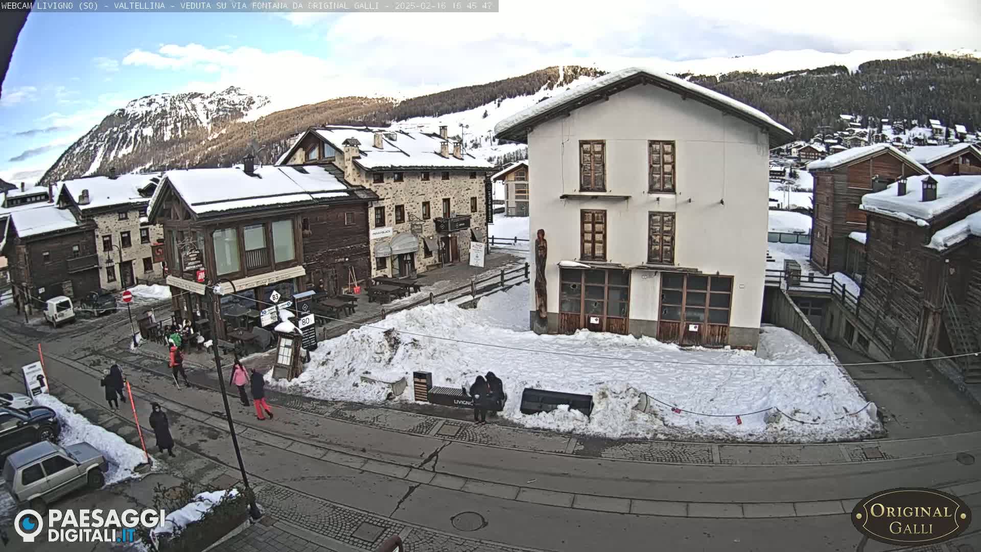 A snow-covered village square with several buildings, some people walking, and a mountainous background under a partly cloudy sky.