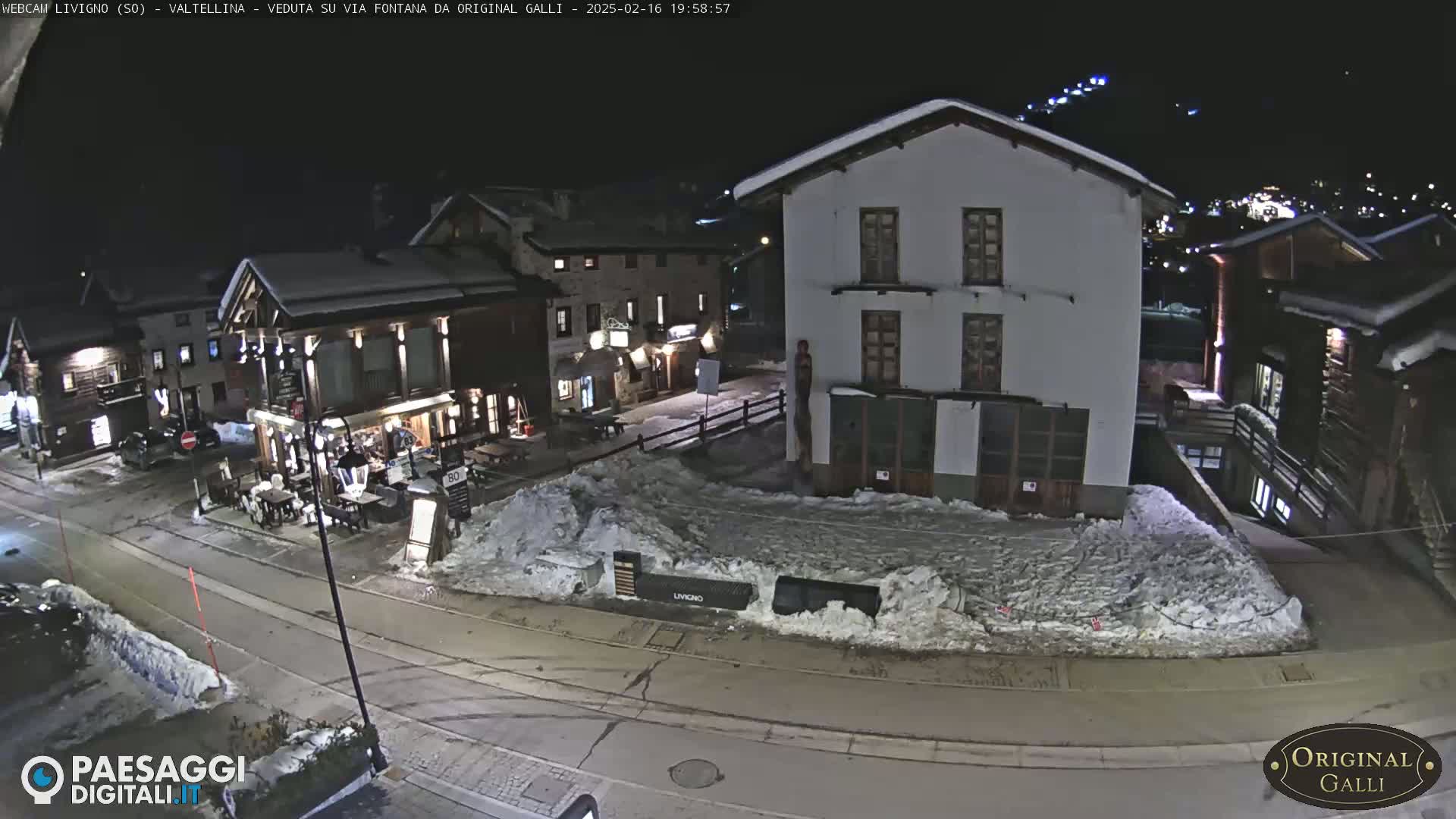 A snow-covered village street at night, illuminated by streetlights, shows several buildings with snow on their roofs.