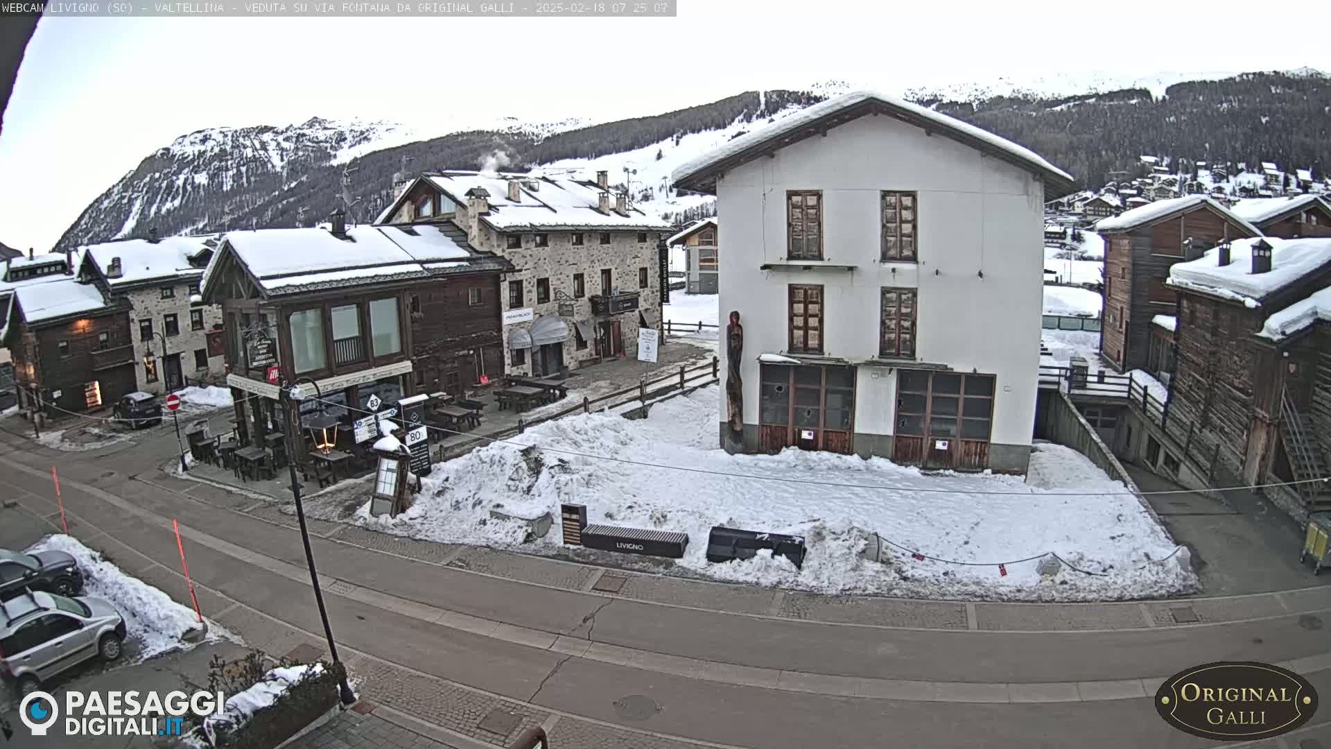 A snow-covered village street scene, with several buildings, some cars parked, and a background of snow-covered mountains under a cloudy sky.