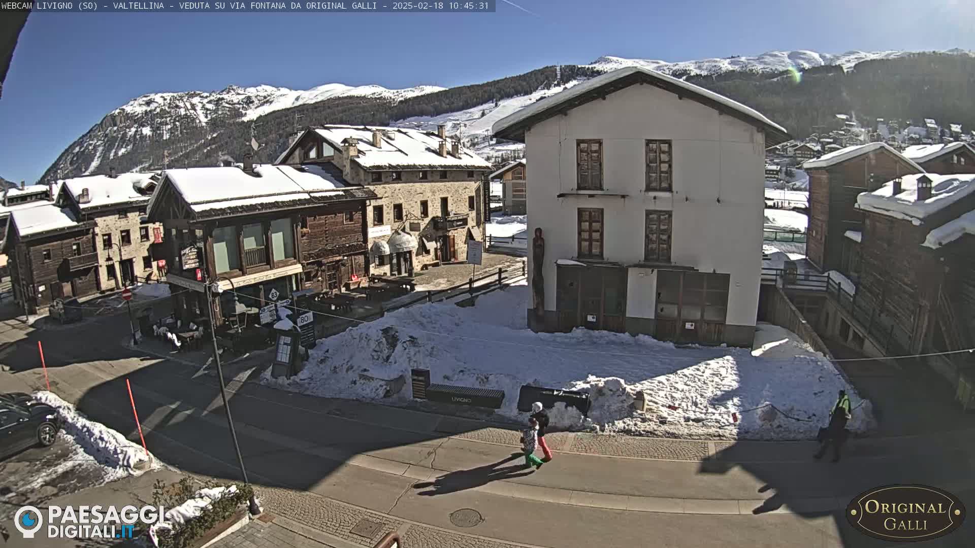 A sunny, snow-covered village scene shows several buildings, a street, and a few pedestrians.