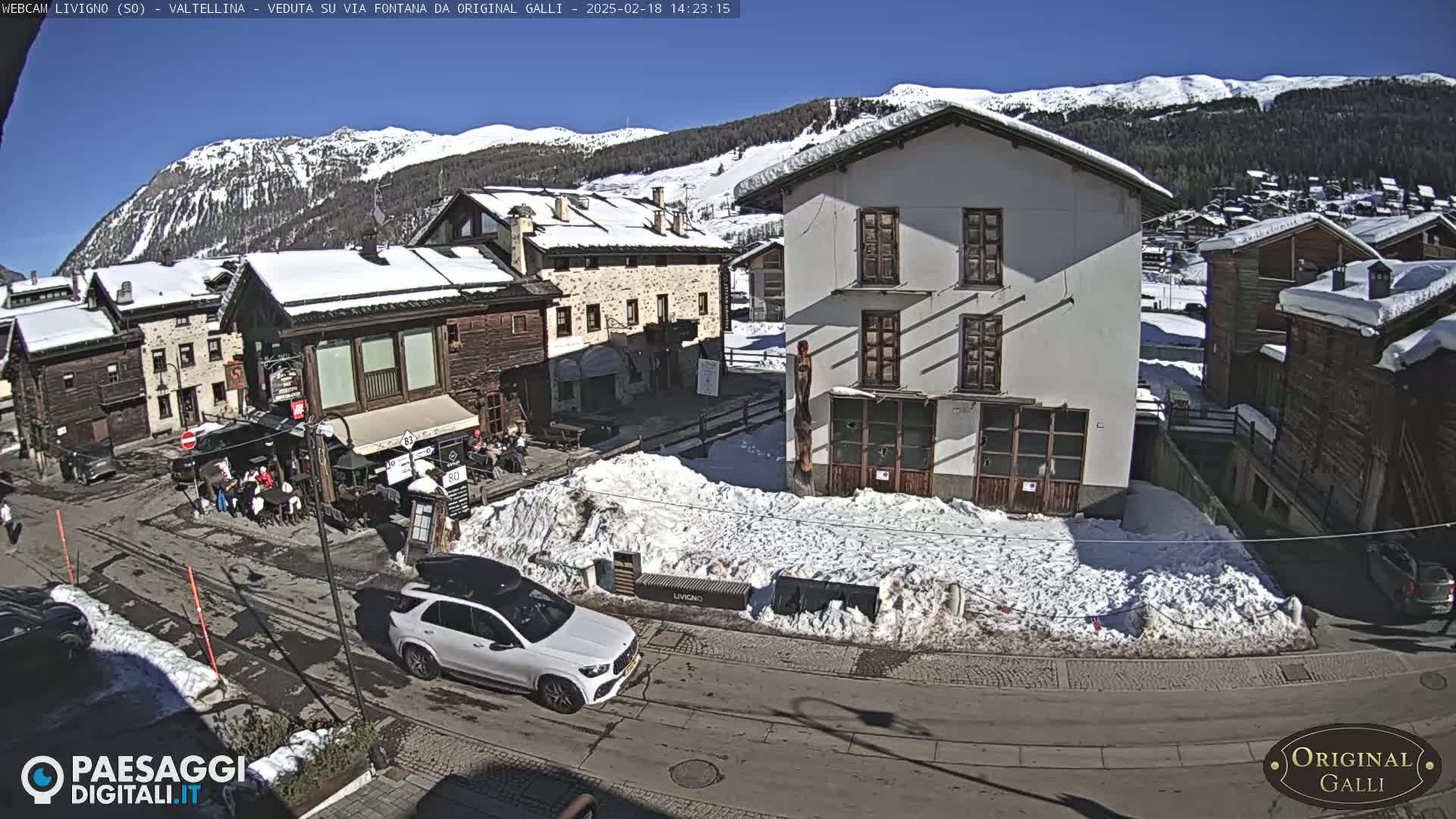 A snow-covered village square with several buildings, a few people, and a car parked on a sunny day with mountains in the background.