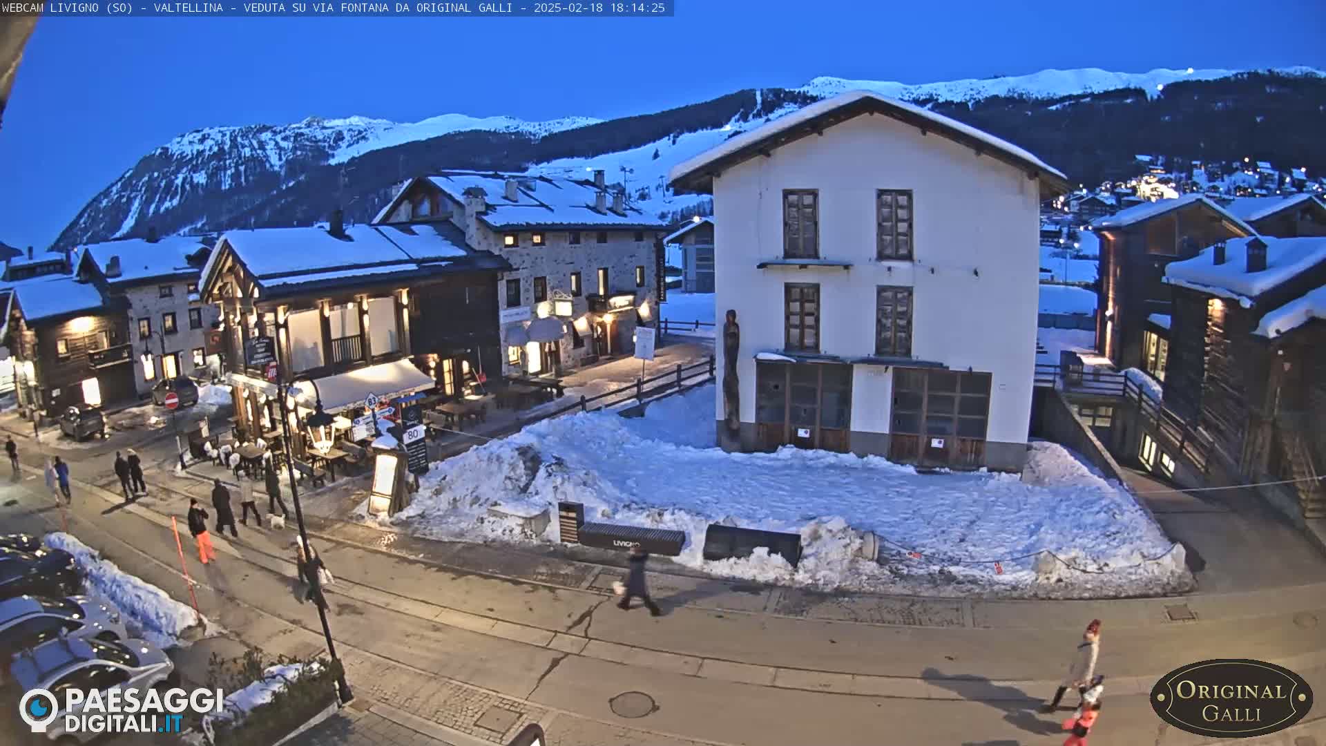 A snow-covered village square at twilight, with people walking on a street lined with buildings, under a clear, dark blue sky and snow-capped mountains in the background.