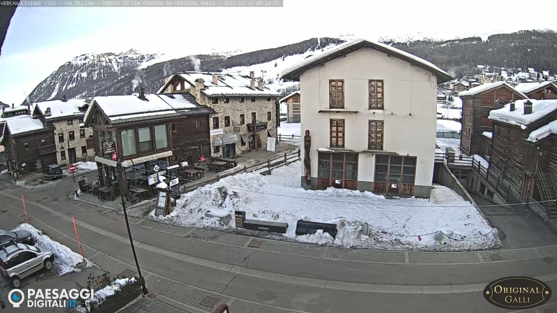 A snow-covered village street scene with several buildings, some cars parked, and a mountain range in the background under a mostly clear sky.