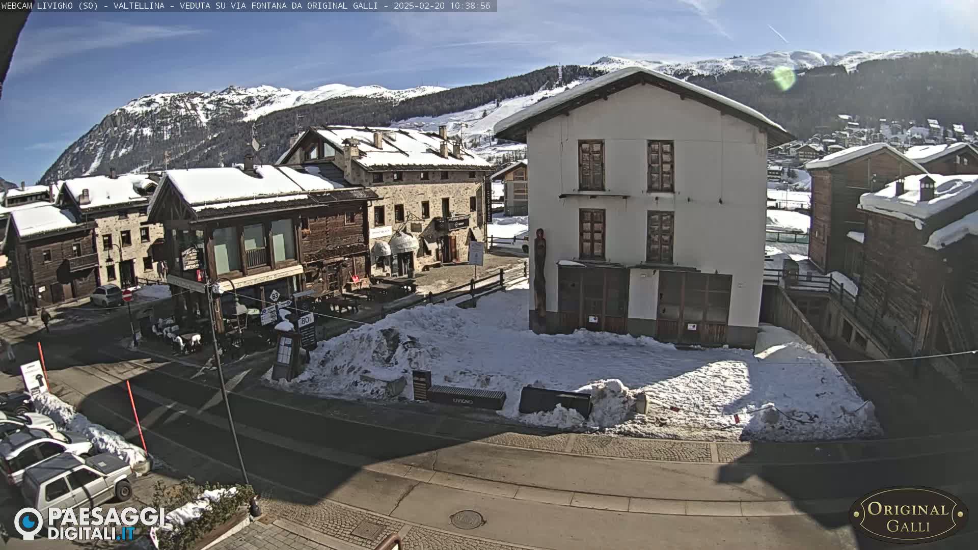A sunny day in a snow-covered village shows several buildings, a street, and cars parked along the side of the road with mountains in the background.