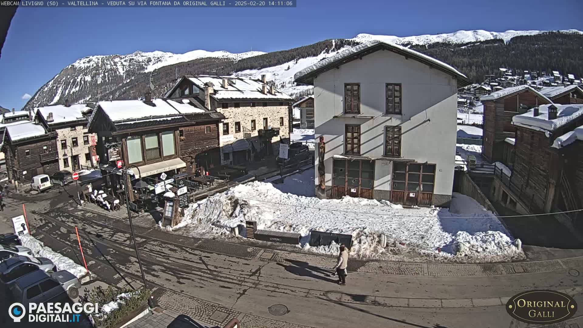 A snow-covered village street scene with buildings, a person walking, and mountains in the background under a sunny sky.