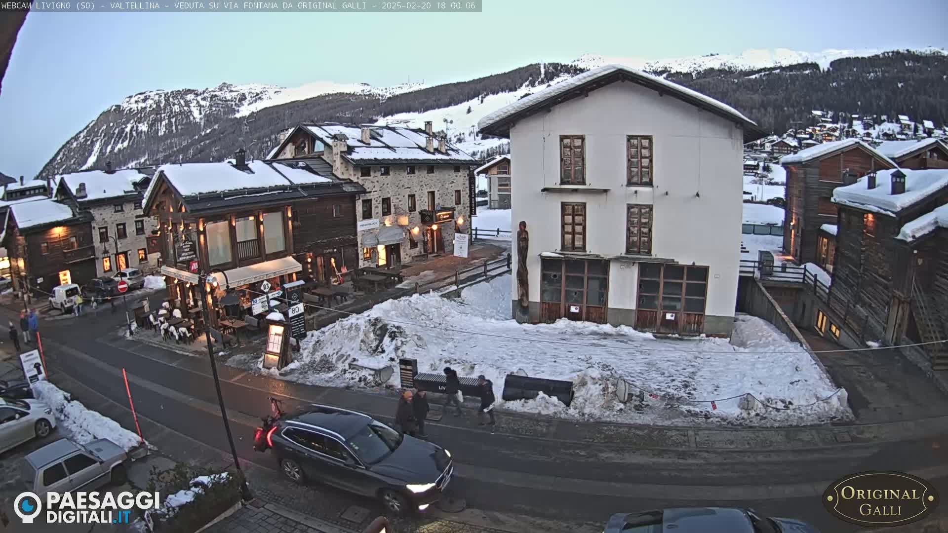 A snowy village square in the mountains, with several buildings and cars visible, under a twilight sky.