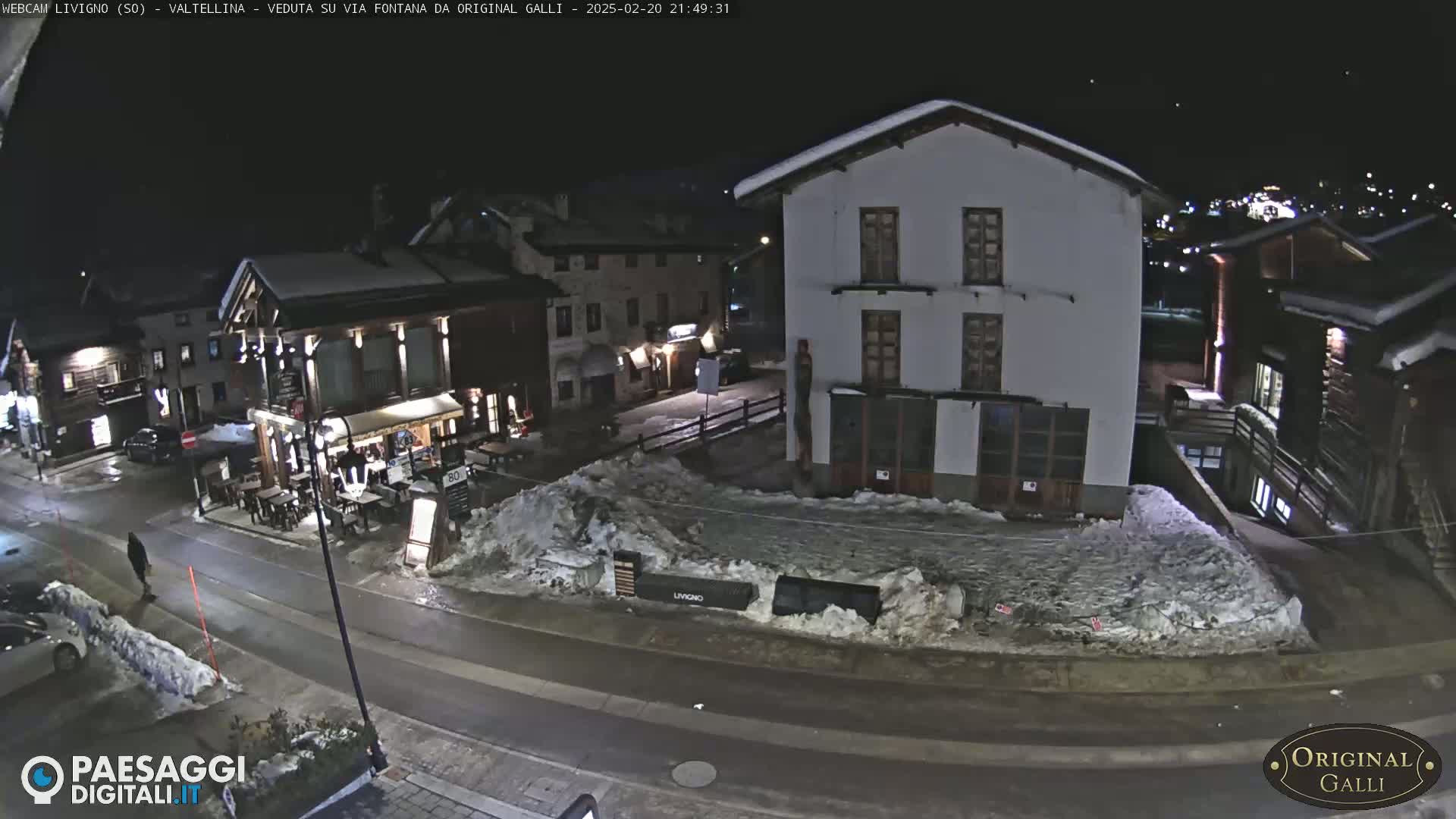 A snow-covered street scene at night shows several buildings, one with outdoor seating, and a pedestrian walking.