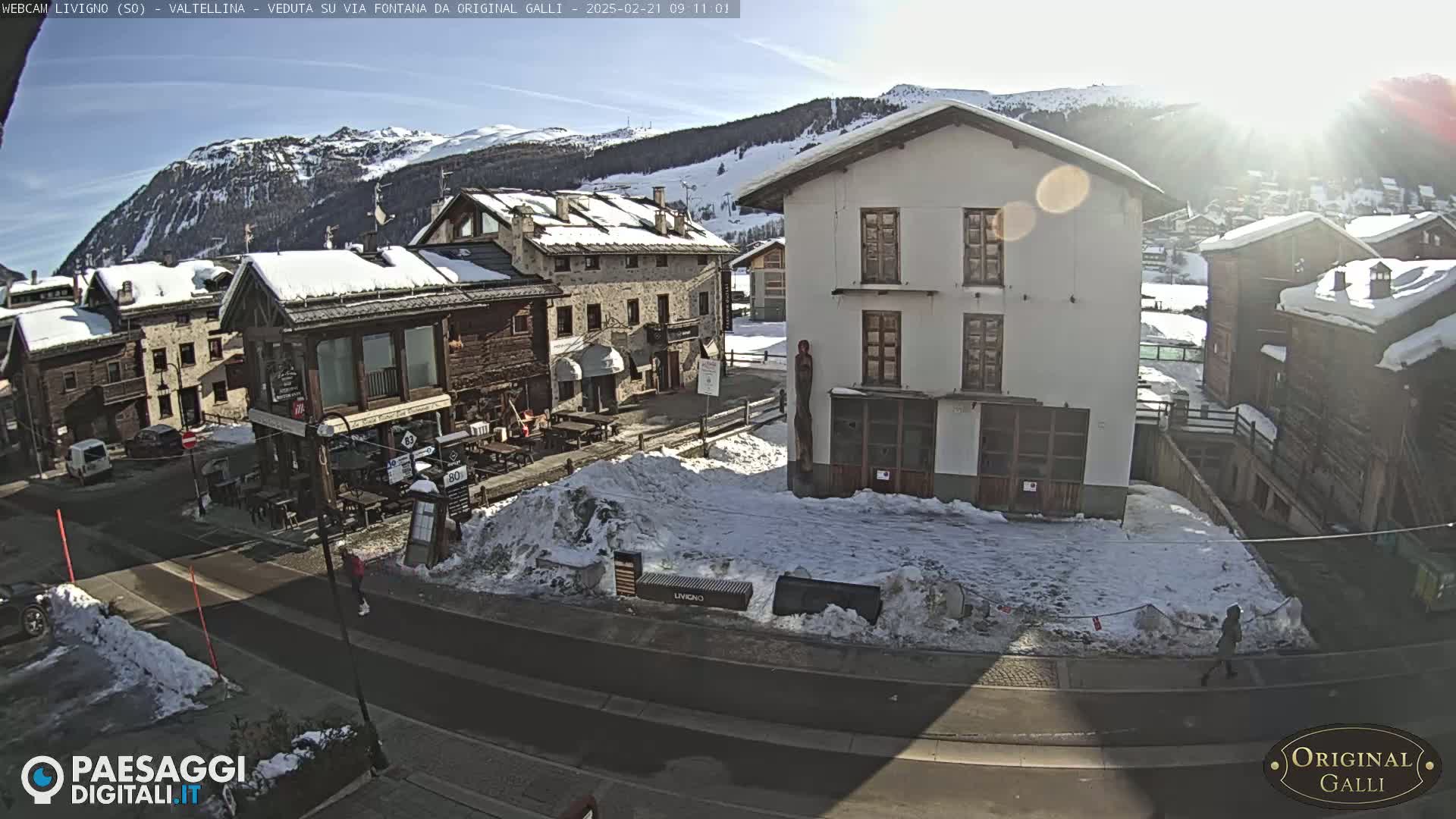 A sunny, snow-covered village street scene in a mountainous area, showing several buildings and a few pedestrians.