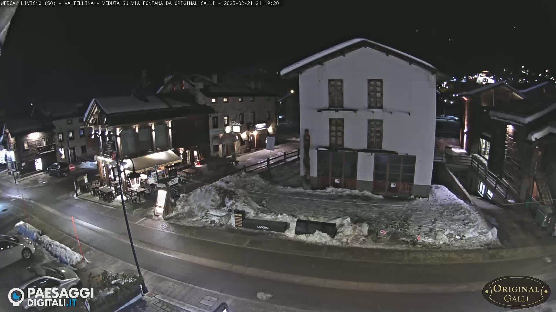 A snow-covered village street at night, with several buildings illuminated, including a restaurant with outdoor seating.