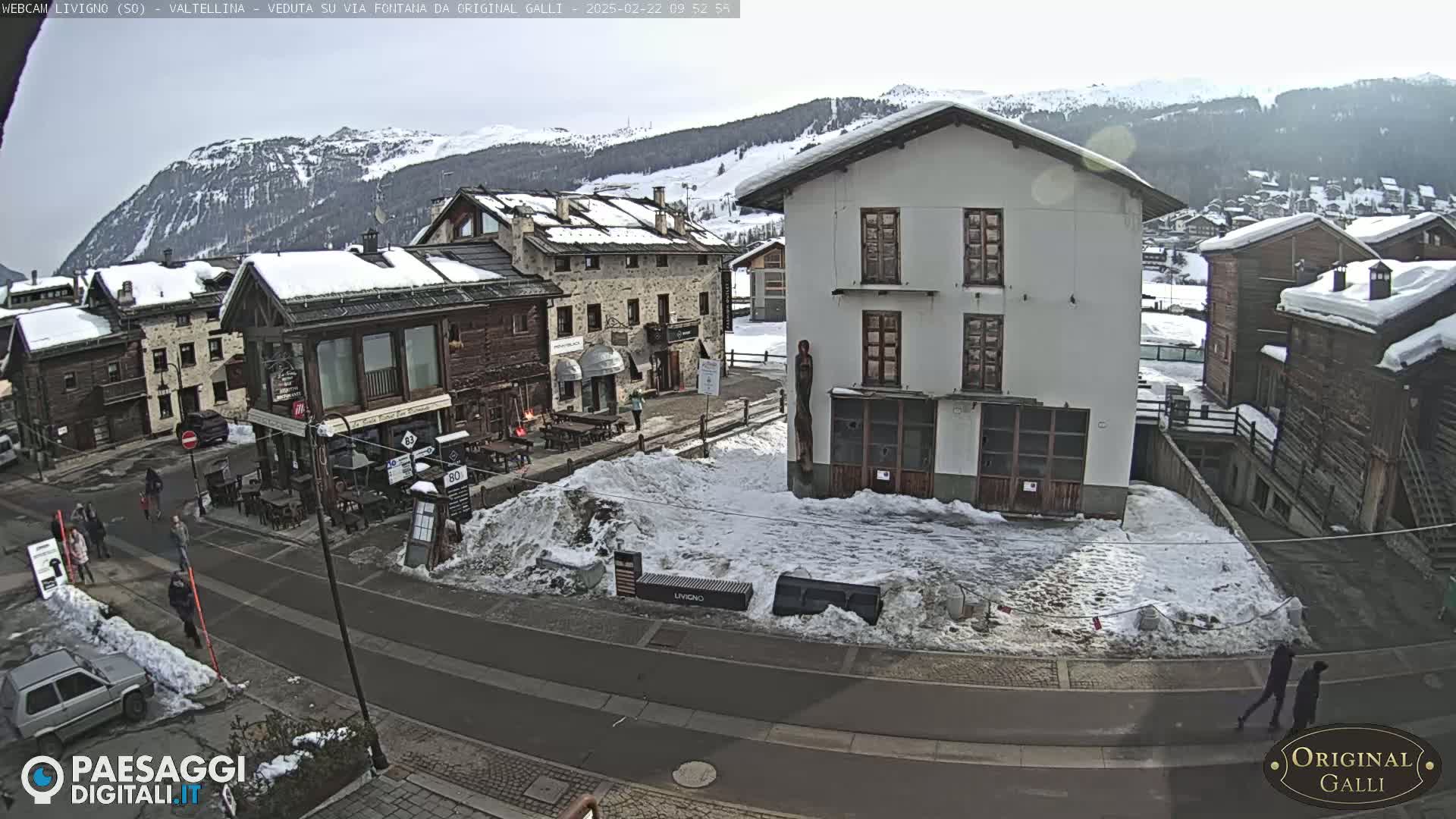 A snow-covered village square with several buildings, some of which are wooden, is seen on a partly cloudy day with mountains in the background.