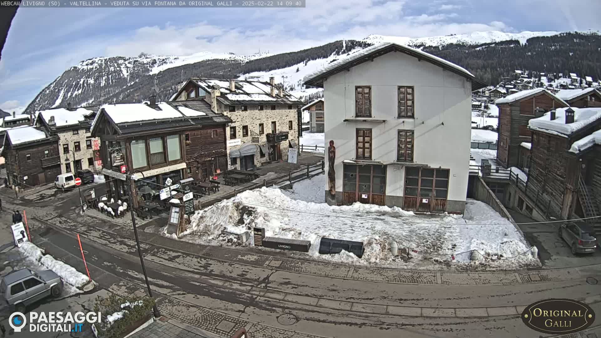 A snow-covered town square, with several buildings and cars, is viewed under a partly sunny sky against a backdrop of snow-capped mountains.