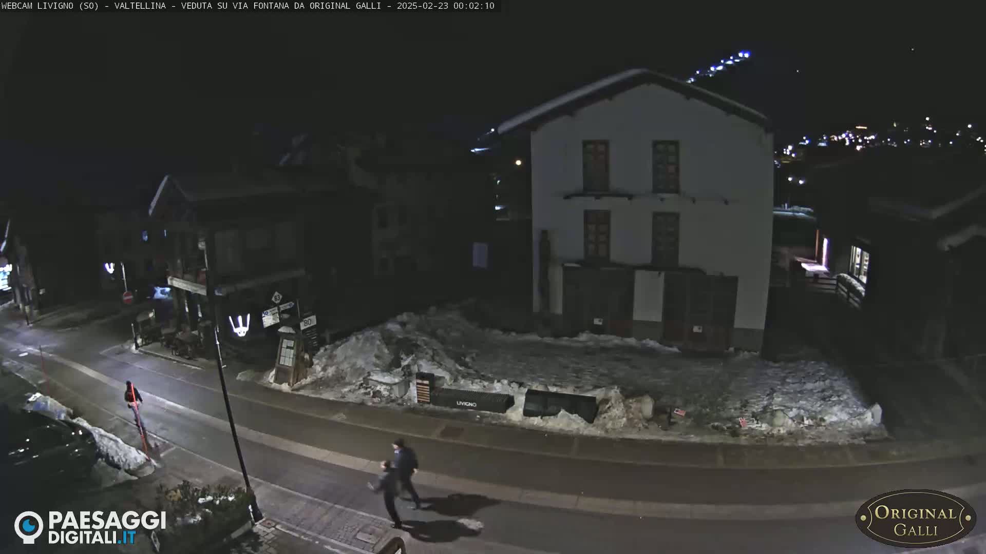 A nighttime street scene shows two people walking on a snow-covered road past buildings under a dark sky.