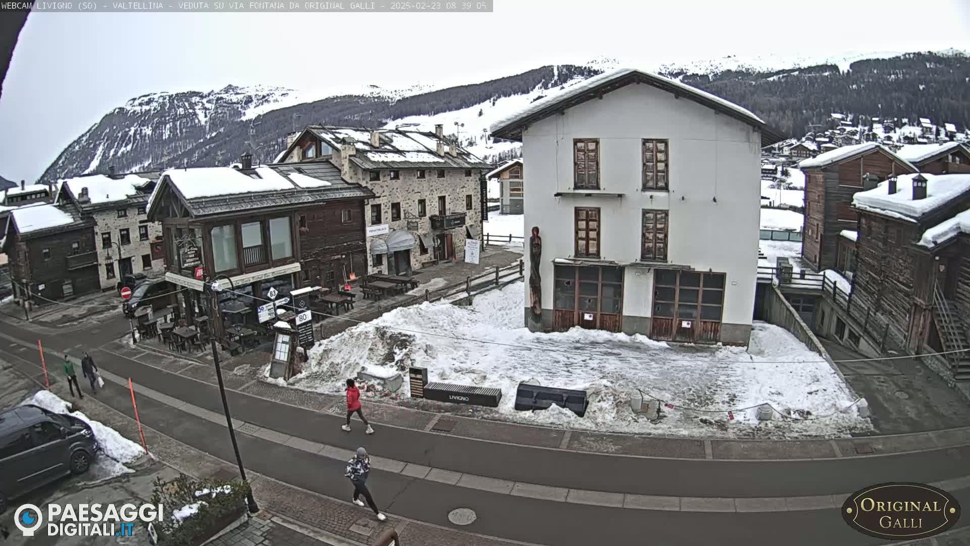 A snow-covered village street scene with several buildings, some pedestrians, and a partly cloudy sky.