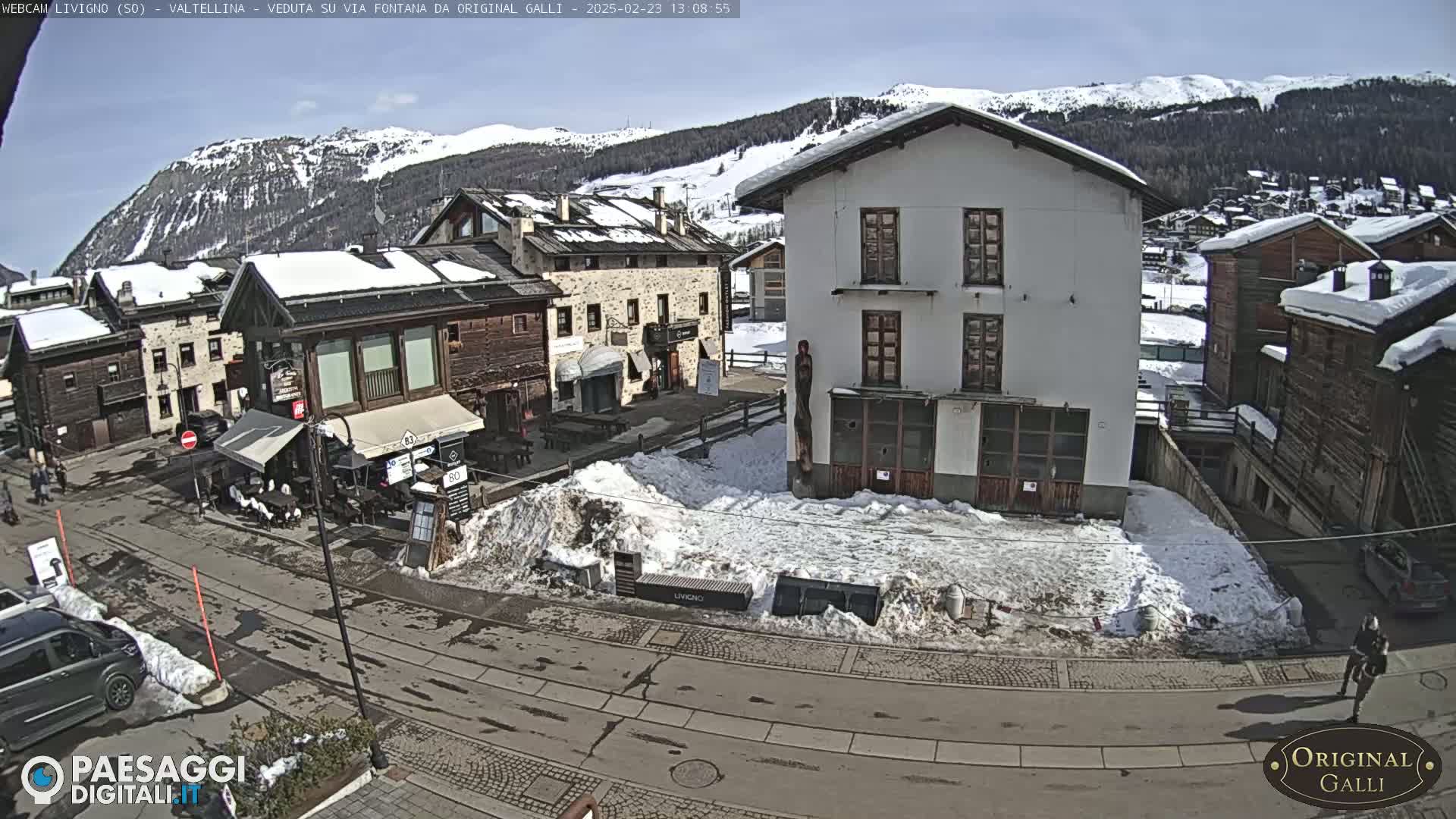 A sunny day in a snow-dusted village shows several buildings, a street, and some parked cars.