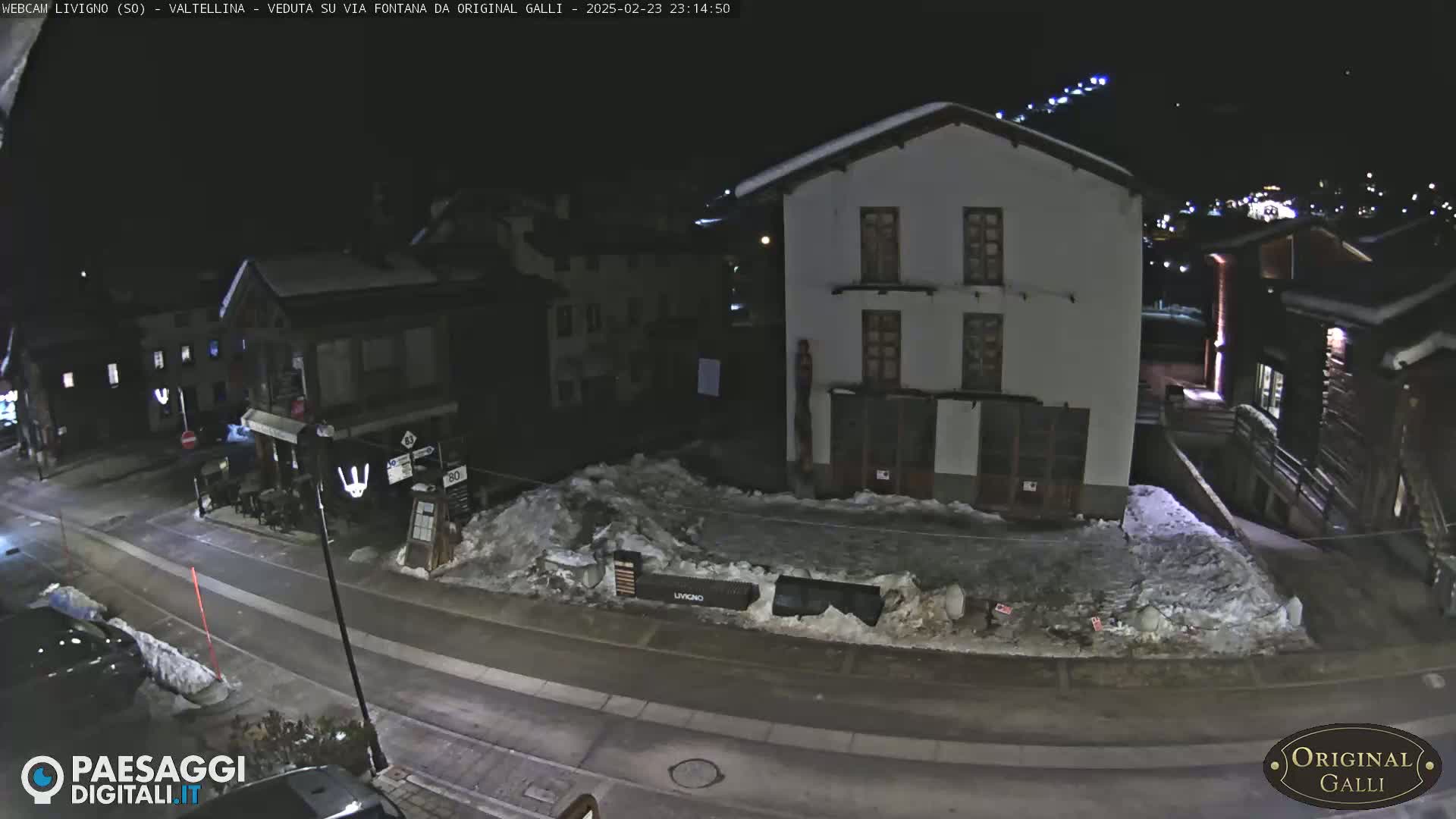 A nighttime street scene in a snow-covered village shows several buildings, a road, and piles of snow.