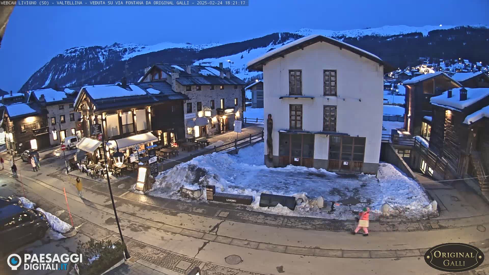 A snow-covered village square at dusk, with several buildings, some with lights on, and a few people walking around.