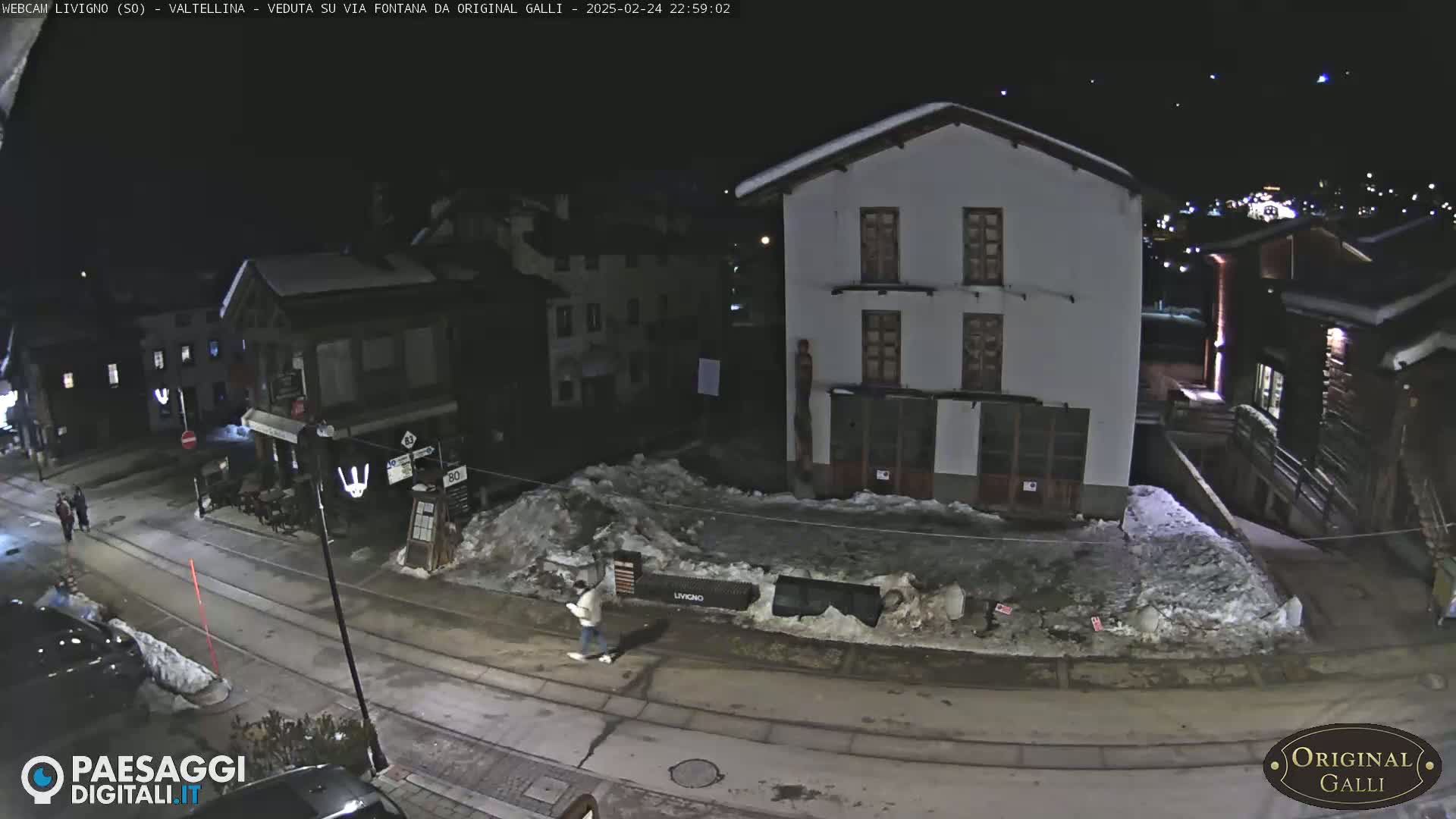 A nighttime street scene in a snow-covered town shows a person walking past buildings under a clear, dark sky.