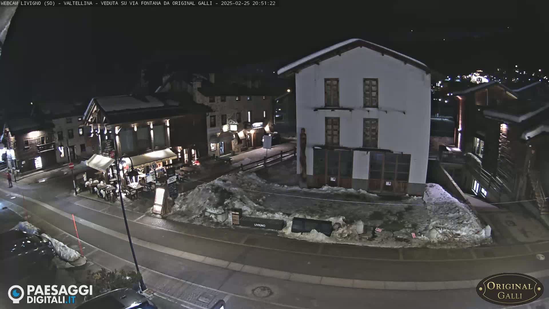 A snow-covered town square at night, with several buildings visible, including a restaurant with outdoor seating.