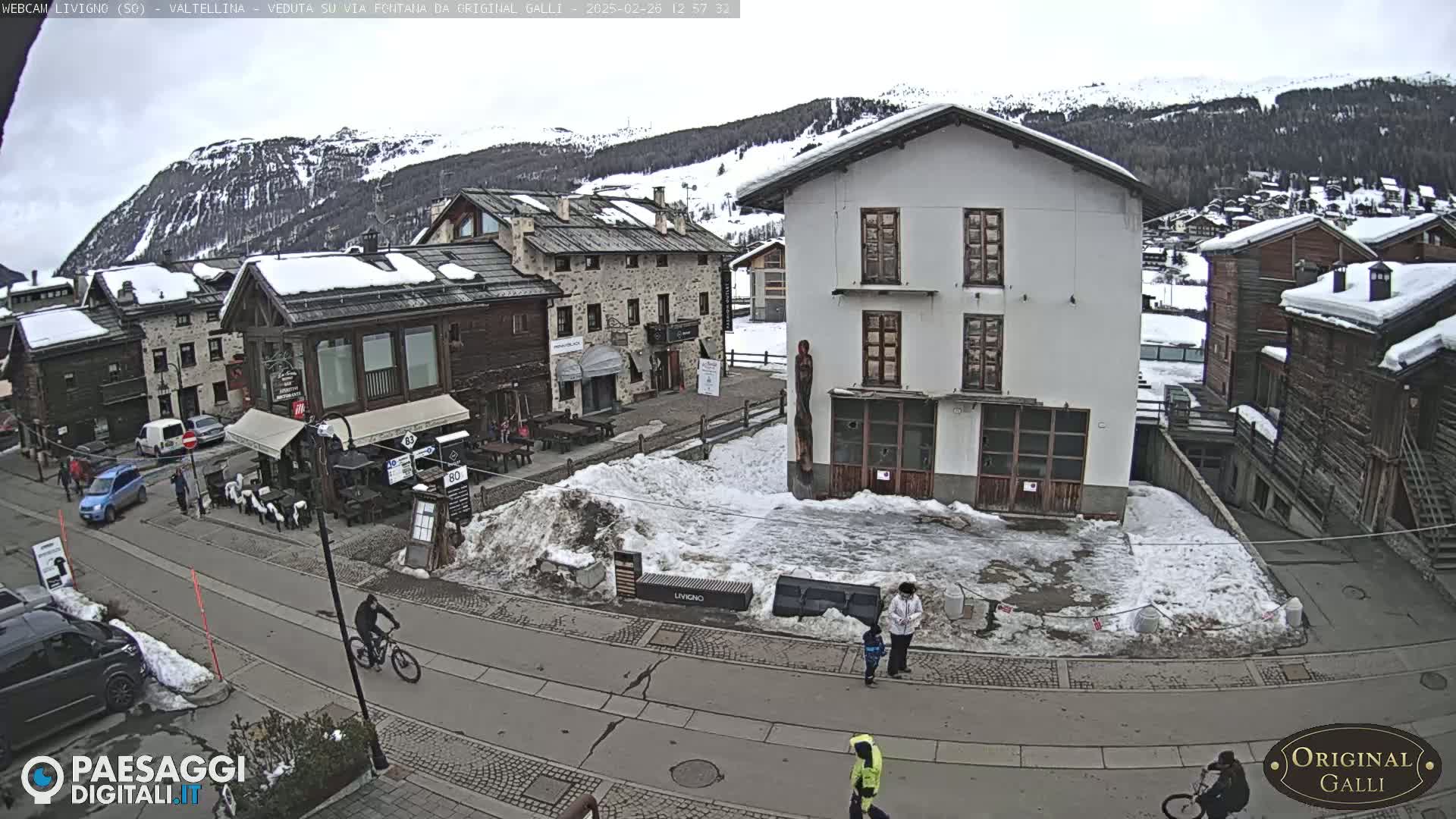 A snowy village square, with several buildings and a few people walking and biking, is seen under an overcast sky.