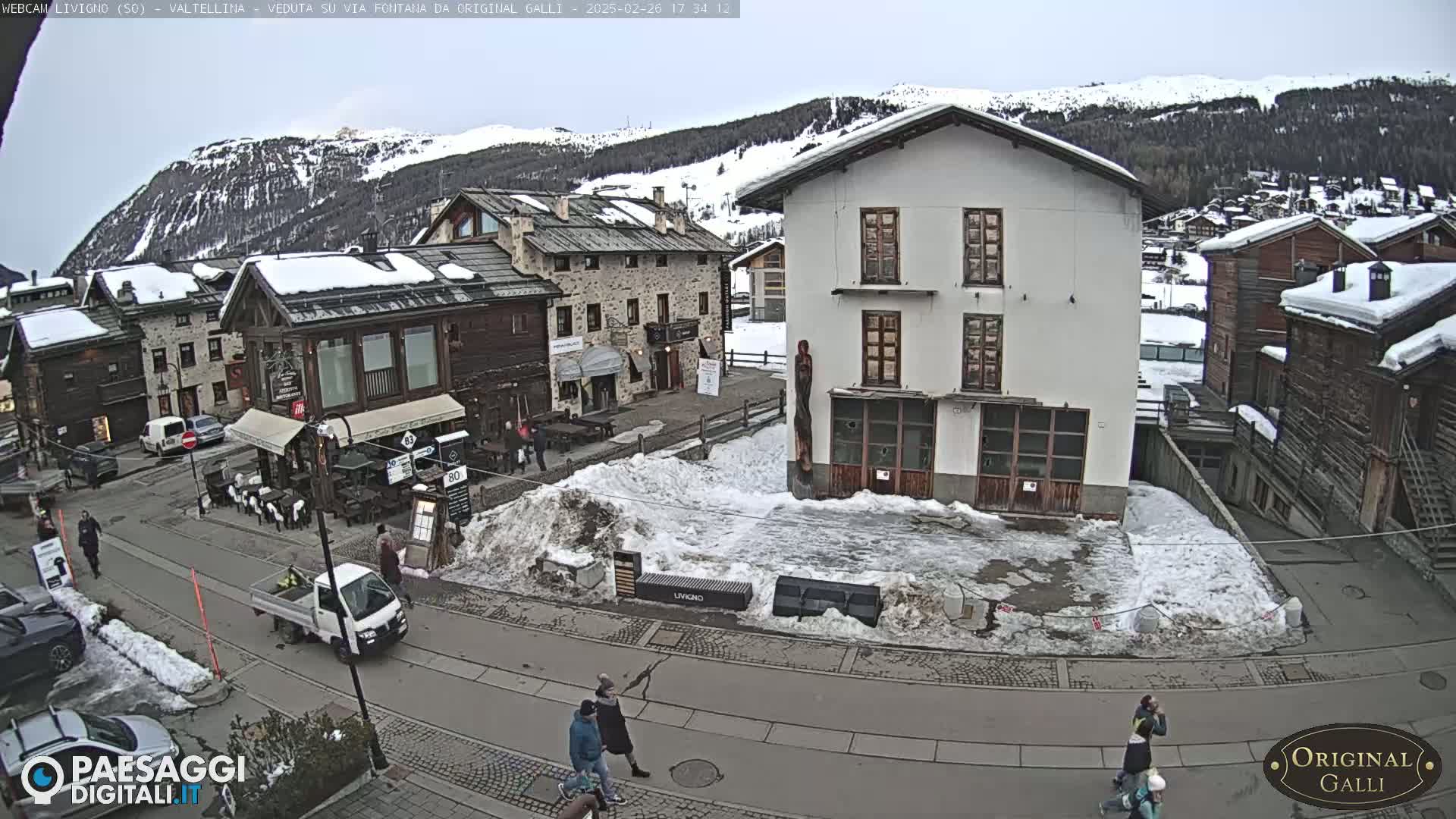 A snow-covered village square, with several buildings and a few people walking around, is seen under an overcast sky.
