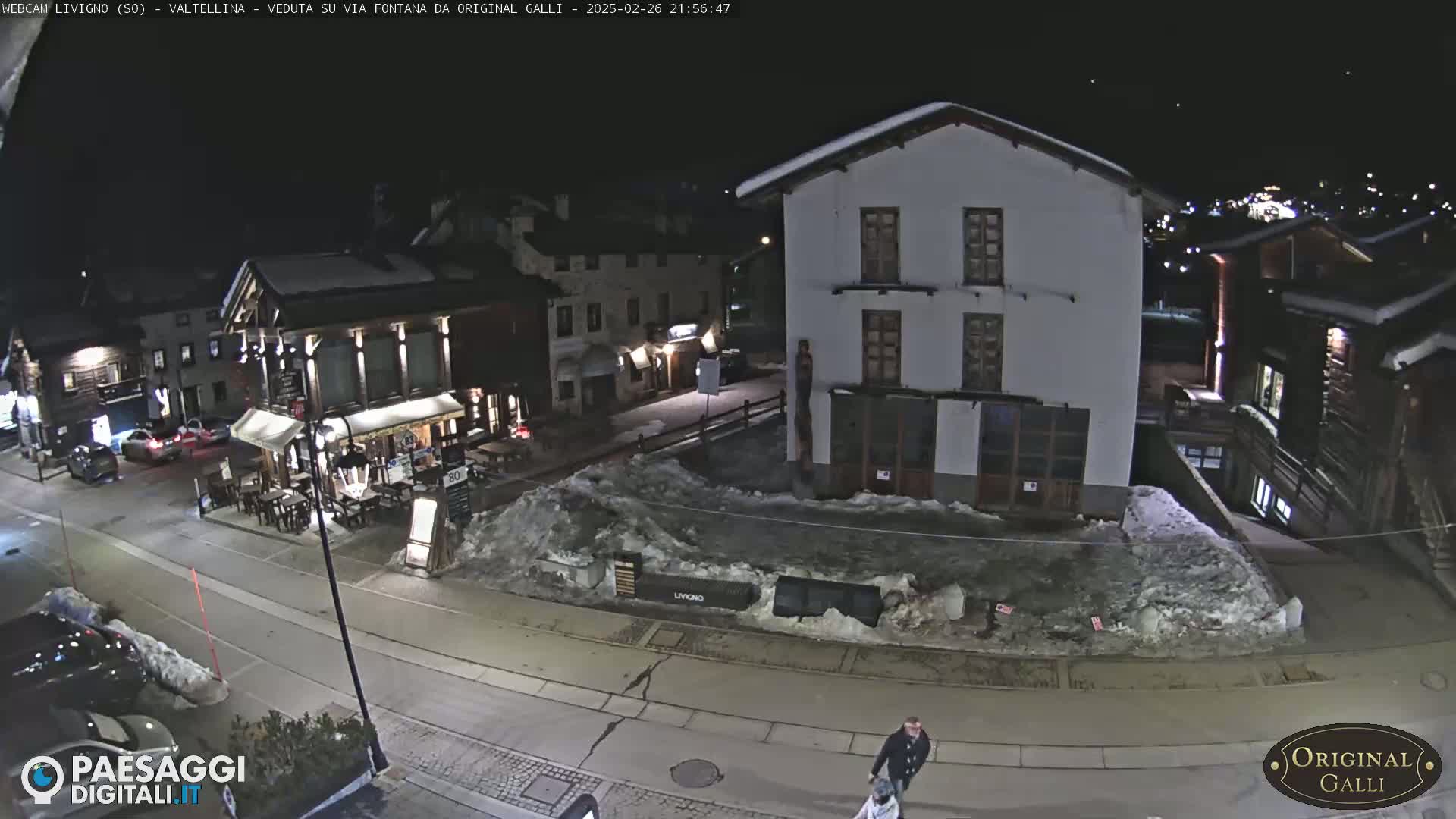 A nighttime street scene in a snow-covered village, showing several buildings, parked cars, and a few pedestrians.