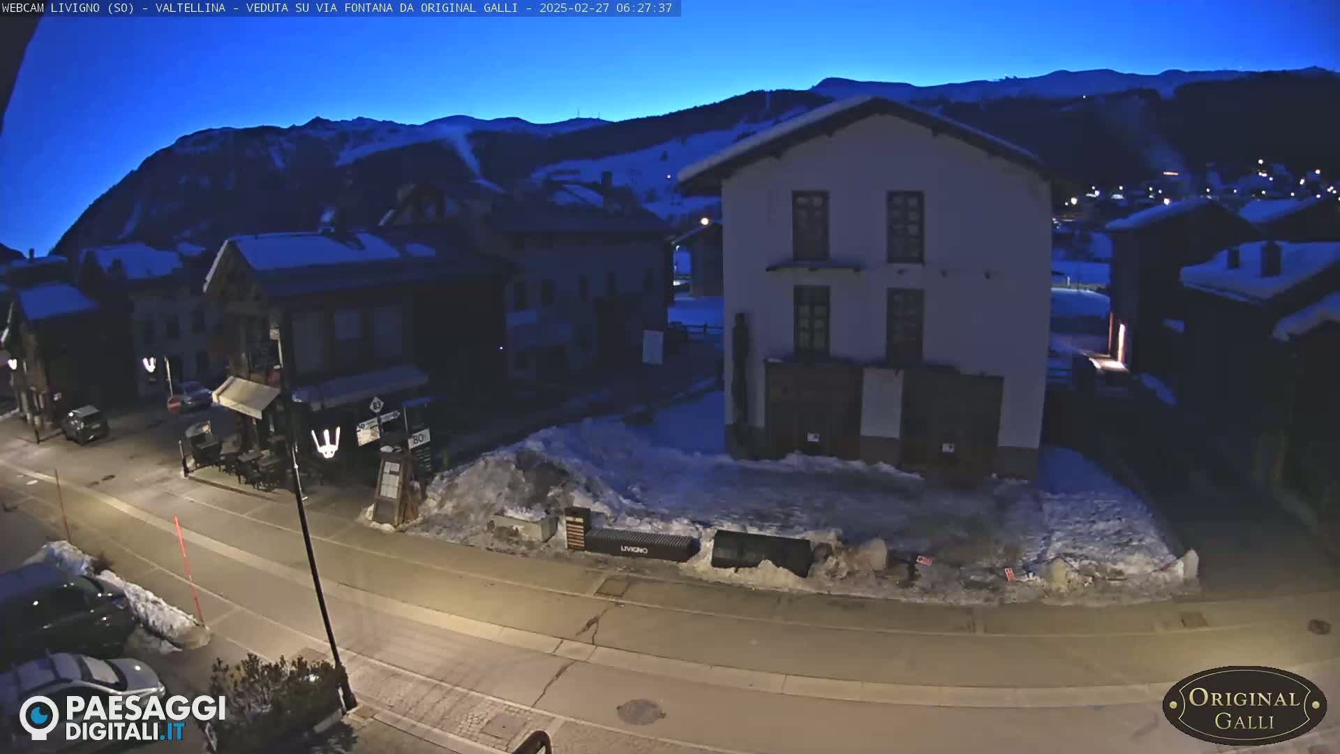 A snow-covered street in a village at twilight, with several buildings visible under a dark blue, pre-dawn sky.