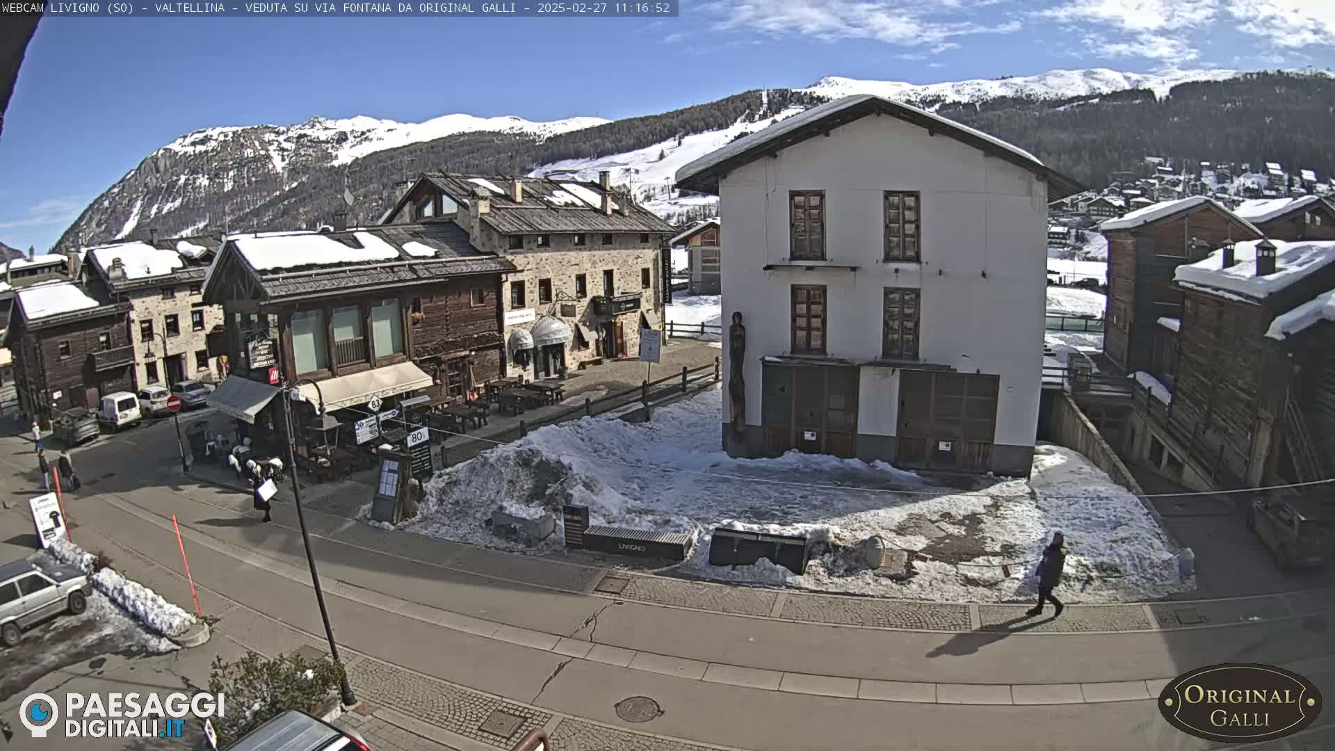A sunny day in a snow-dusted village nestled in a mountain valley shows a street scene with buildings, parked cars, and a few pedestrians.