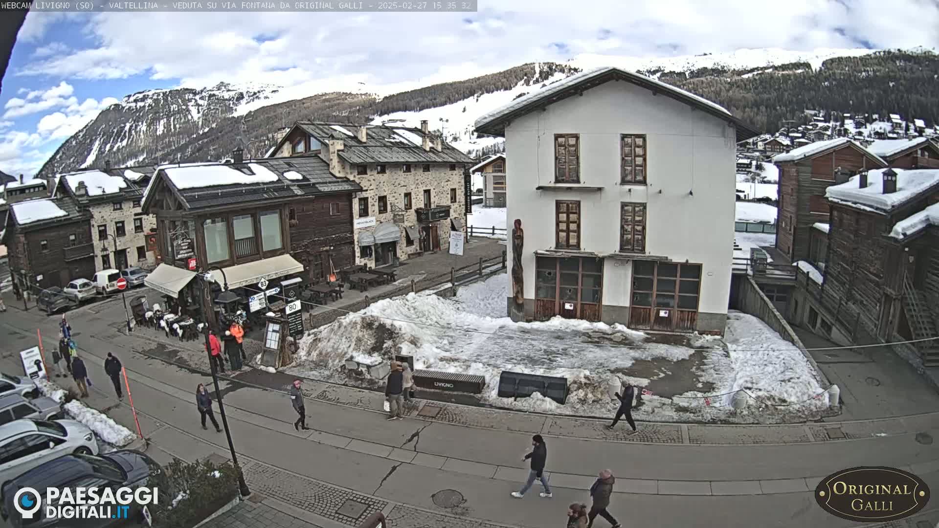 A partly snowy village street scene is shown, with pedestrians walking on a road, buildings lining the street, and snow-covered ground and mountains in the background under a partly cloudy sky.
