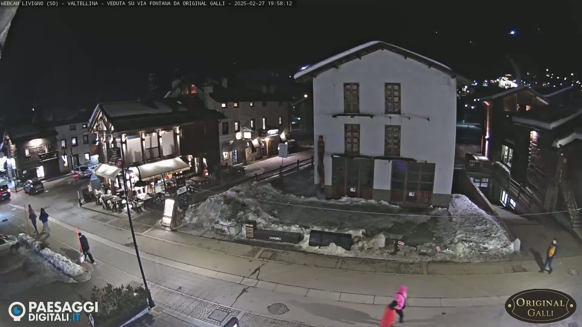 A nighttime street scene in a snow-covered village shows several buildings, a few people walking, and parked cars.