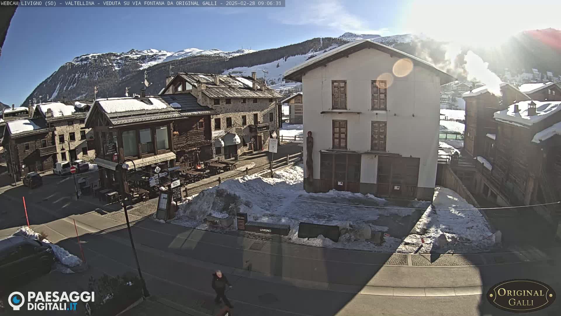 A sunny day in a snow-covered mountain village, showing several buildings and a mostly empty street.