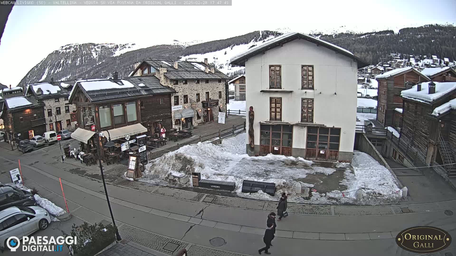 A snow-covered village square with several buildings, some cars parked, and two pedestrians walking, under a mostly cloudy sky.