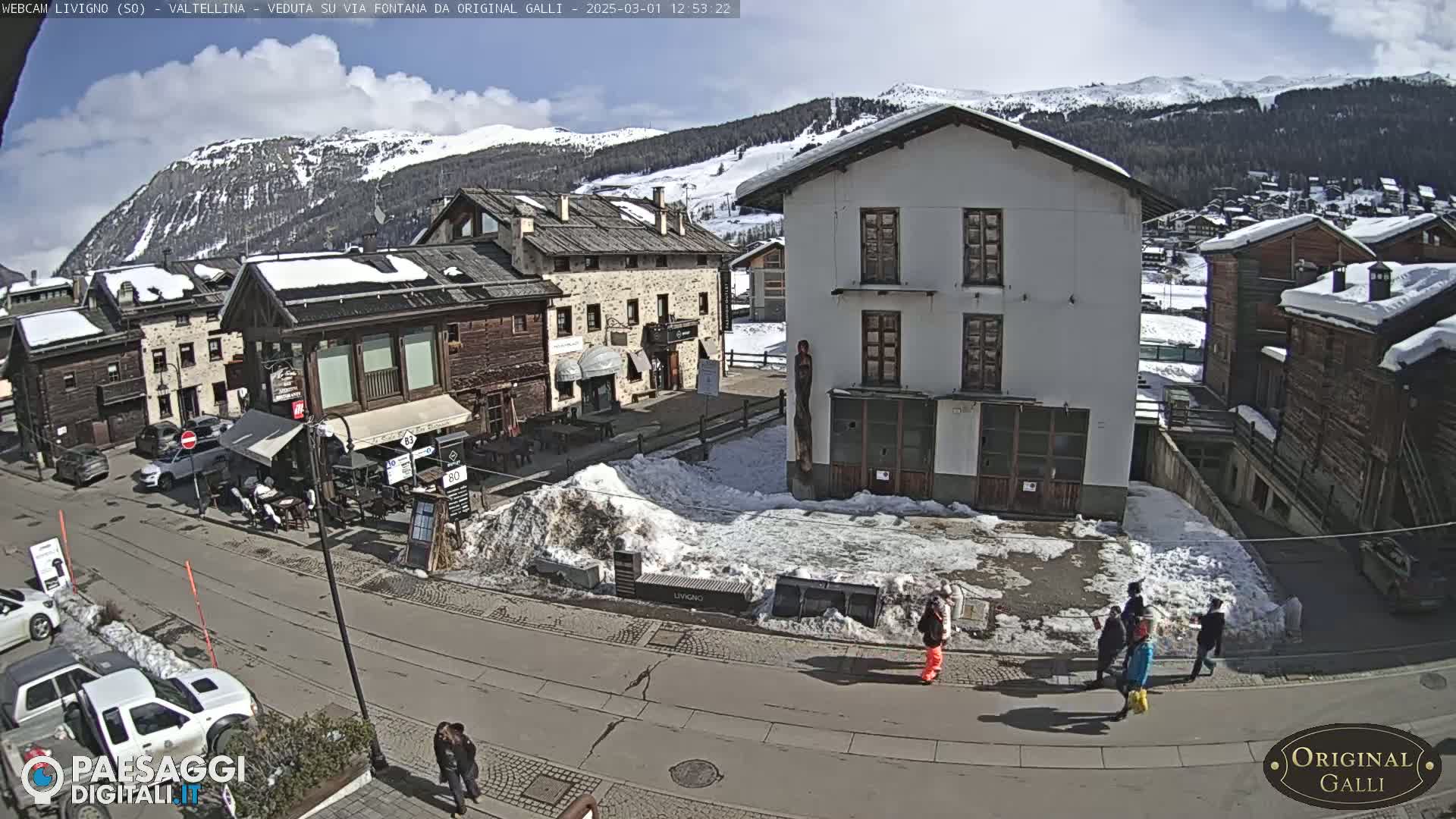 A sunny day in a snow-dusted village nestled in a mountain valley, with several people walking on a street near buildings.
