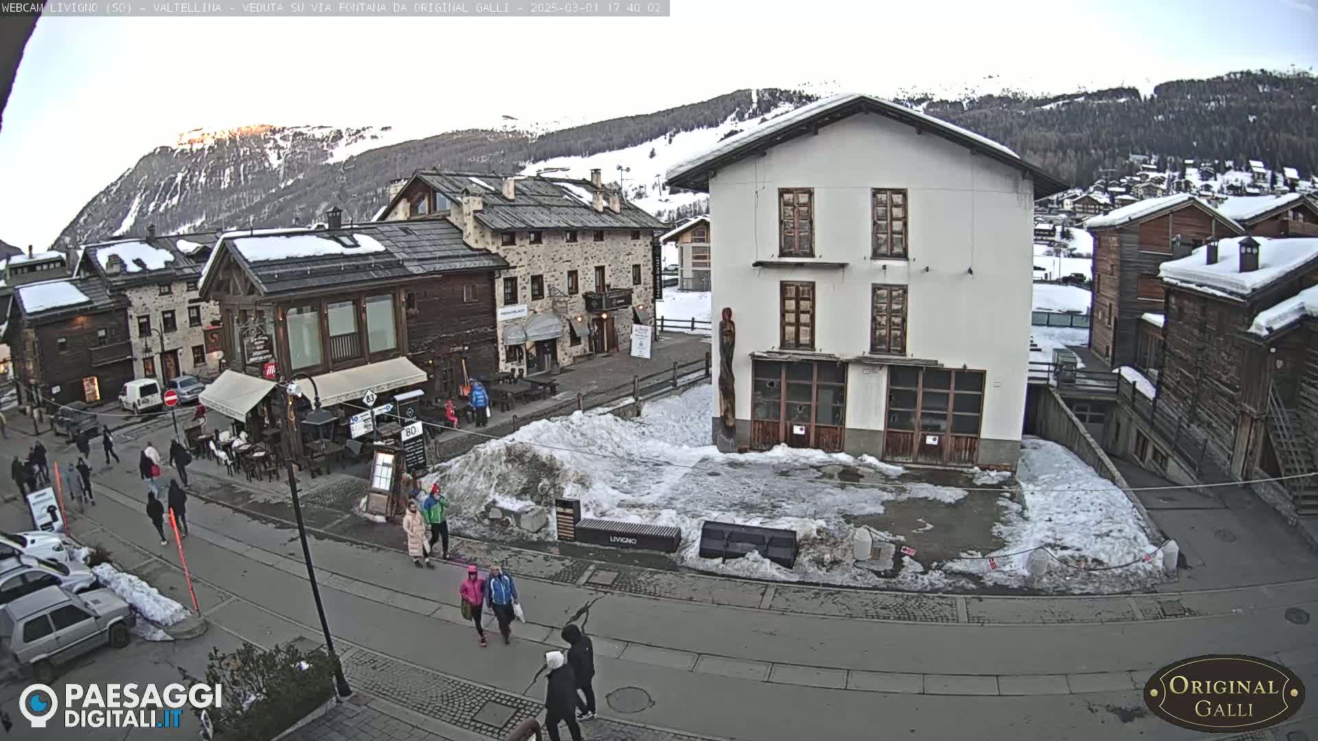 A snowy village square in the mountains, with several pedestrians and parked cars, under a mostly clear sky.