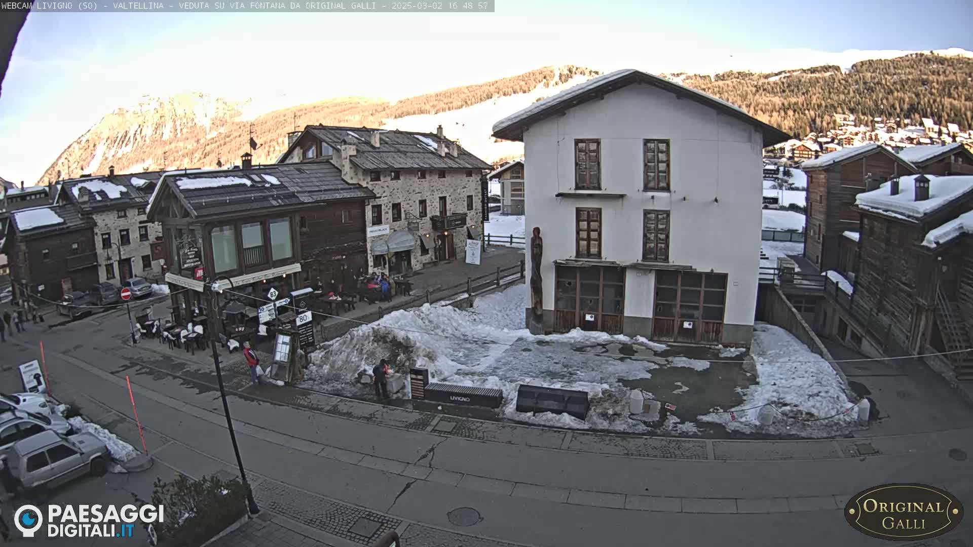 A sunny view of a town square in a snow-dusted mountain village, with several buildings and a few people visible.