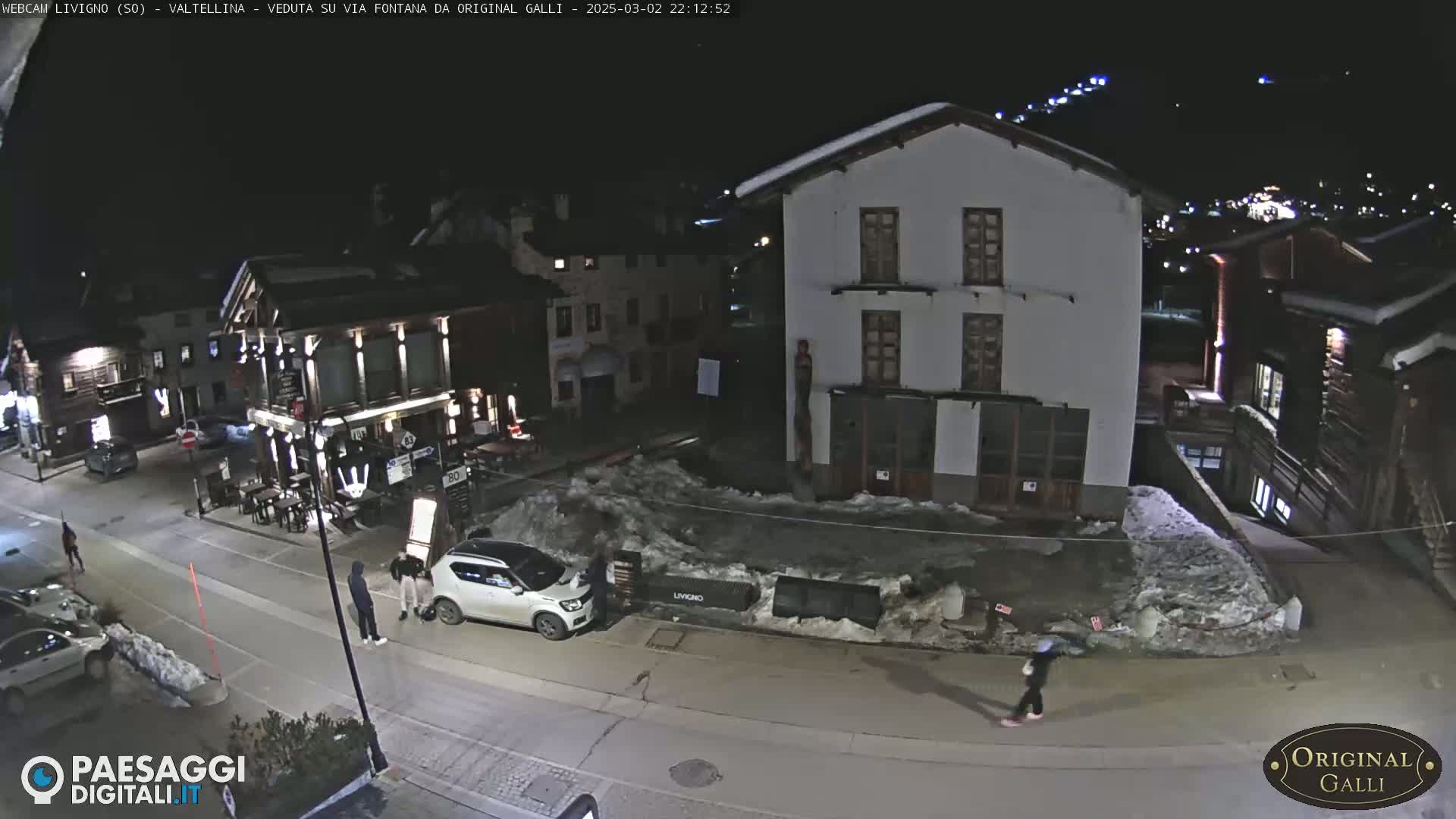 A nighttime street scene in a snow-covered village shows several buildings, a few pedestrians, and parked cars.