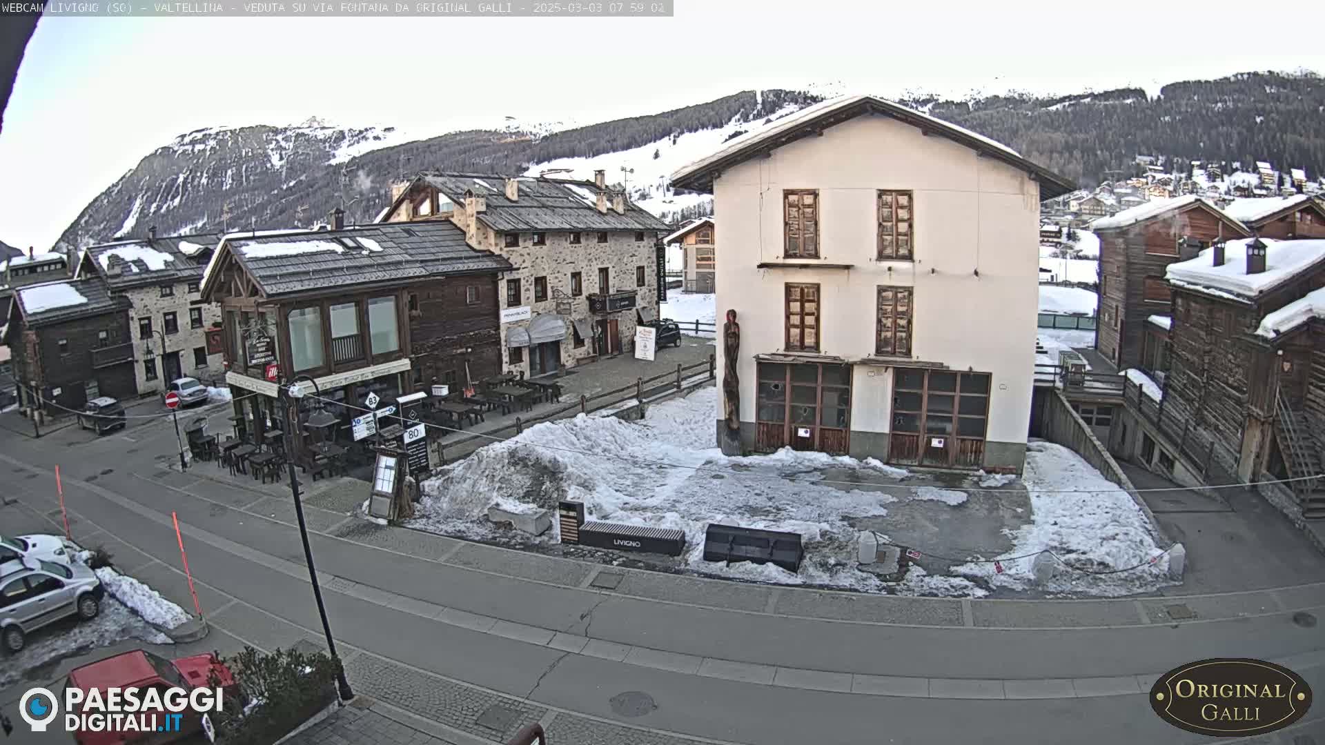 A snow-covered village square with several stone and wood buildings, some with outdoor seating, is visible under a cloudy sky.