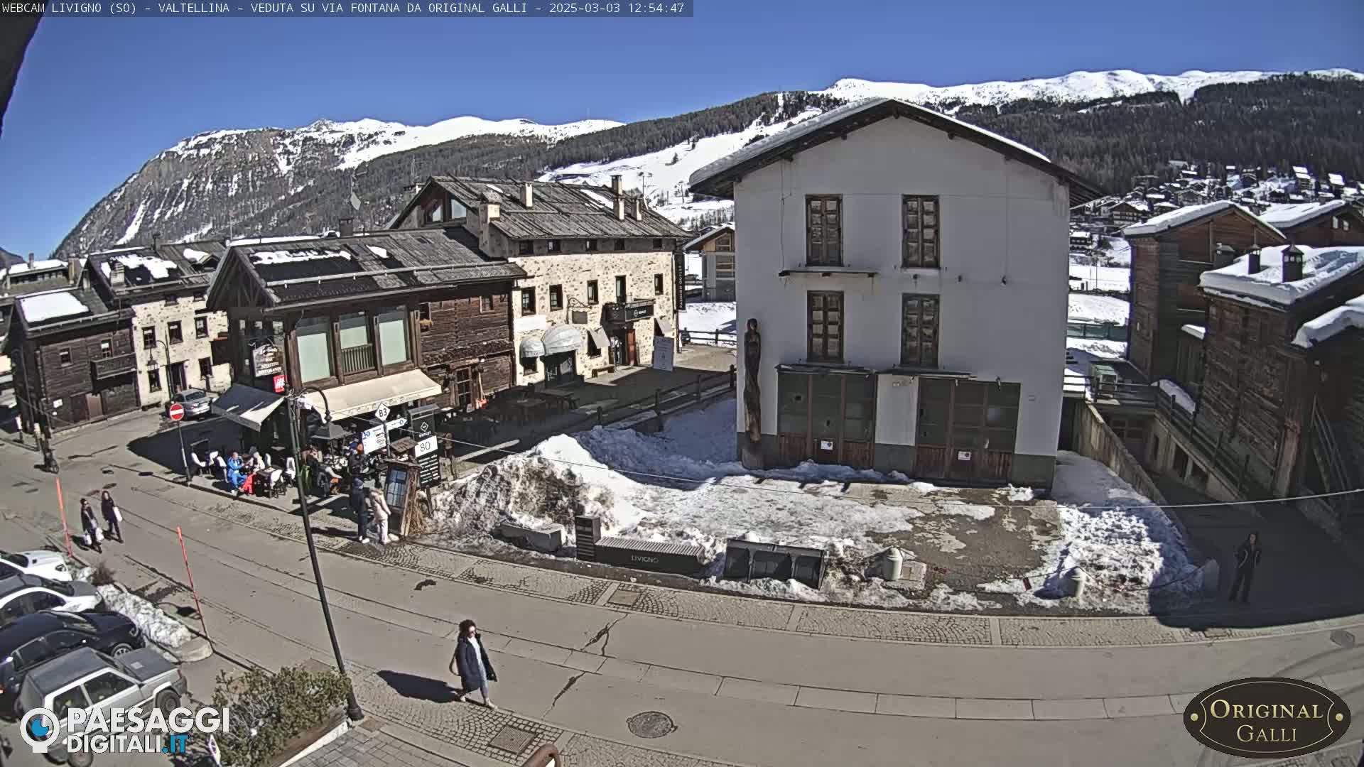 A sunny day in a snow-dusted mountain village shows several buildings surrounding a small town square with people walking and sitting outside cafes.