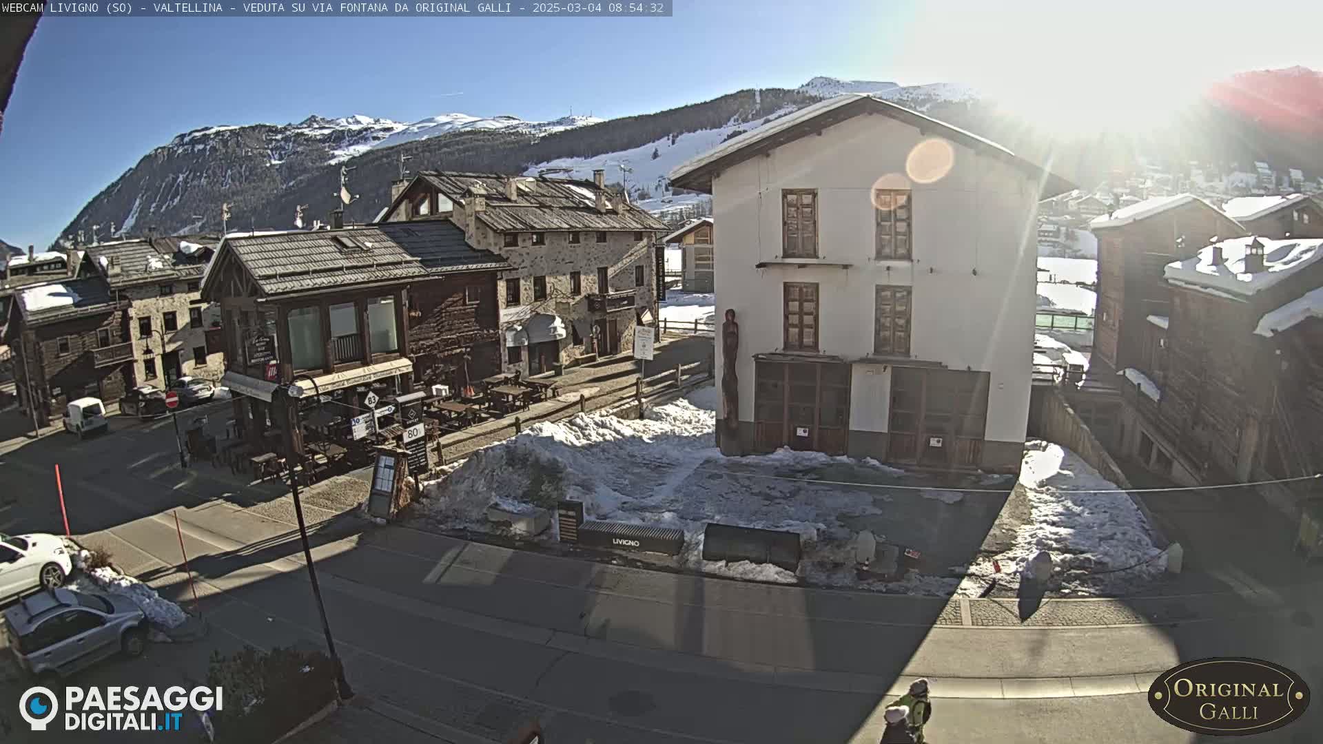 A sunny, snow-dusted village street scene with several buildings, some cars, and patches of snow on the ground.