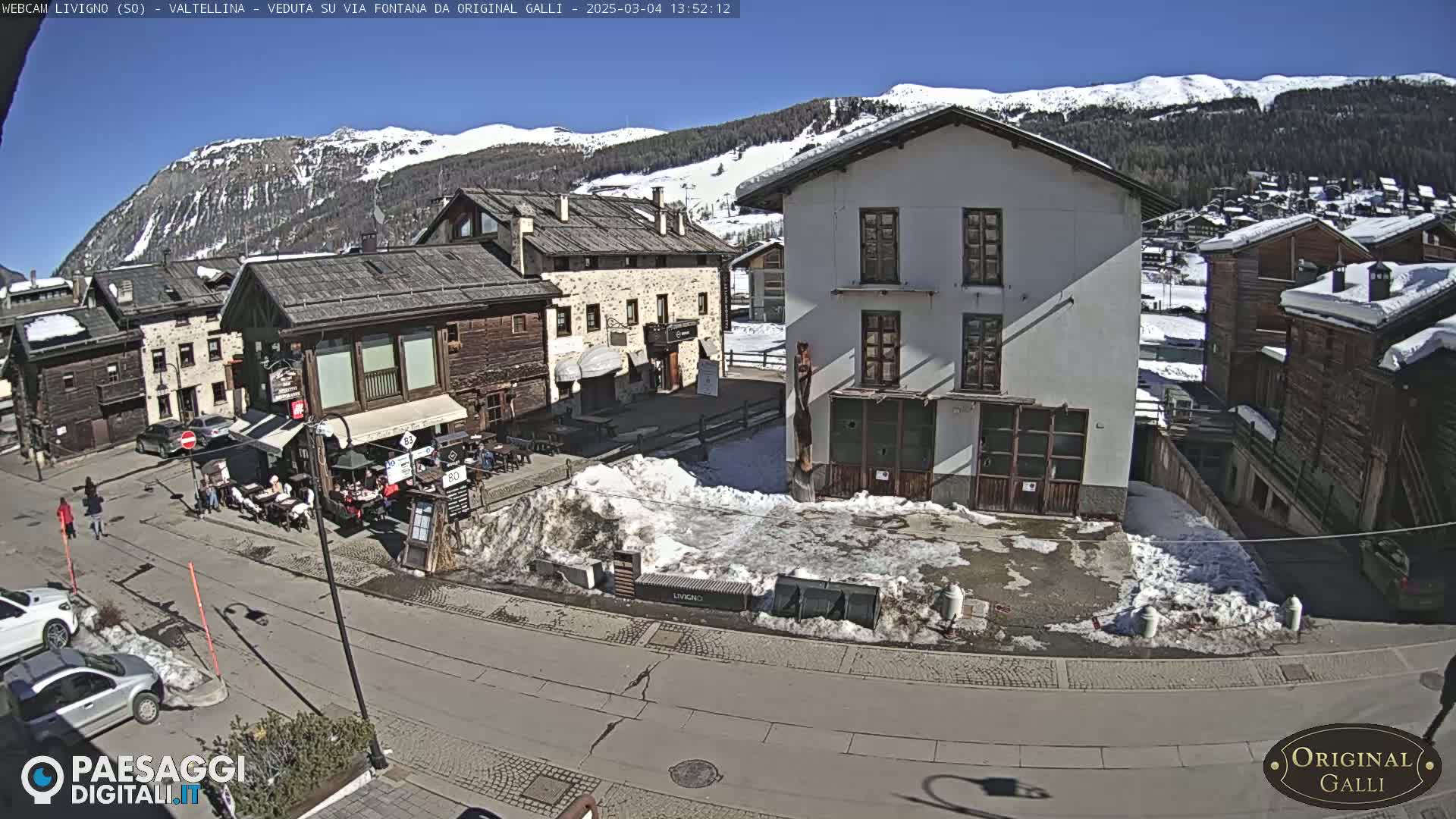 A sunny mountain village square with snow patches, several buildings, and a few people walking.