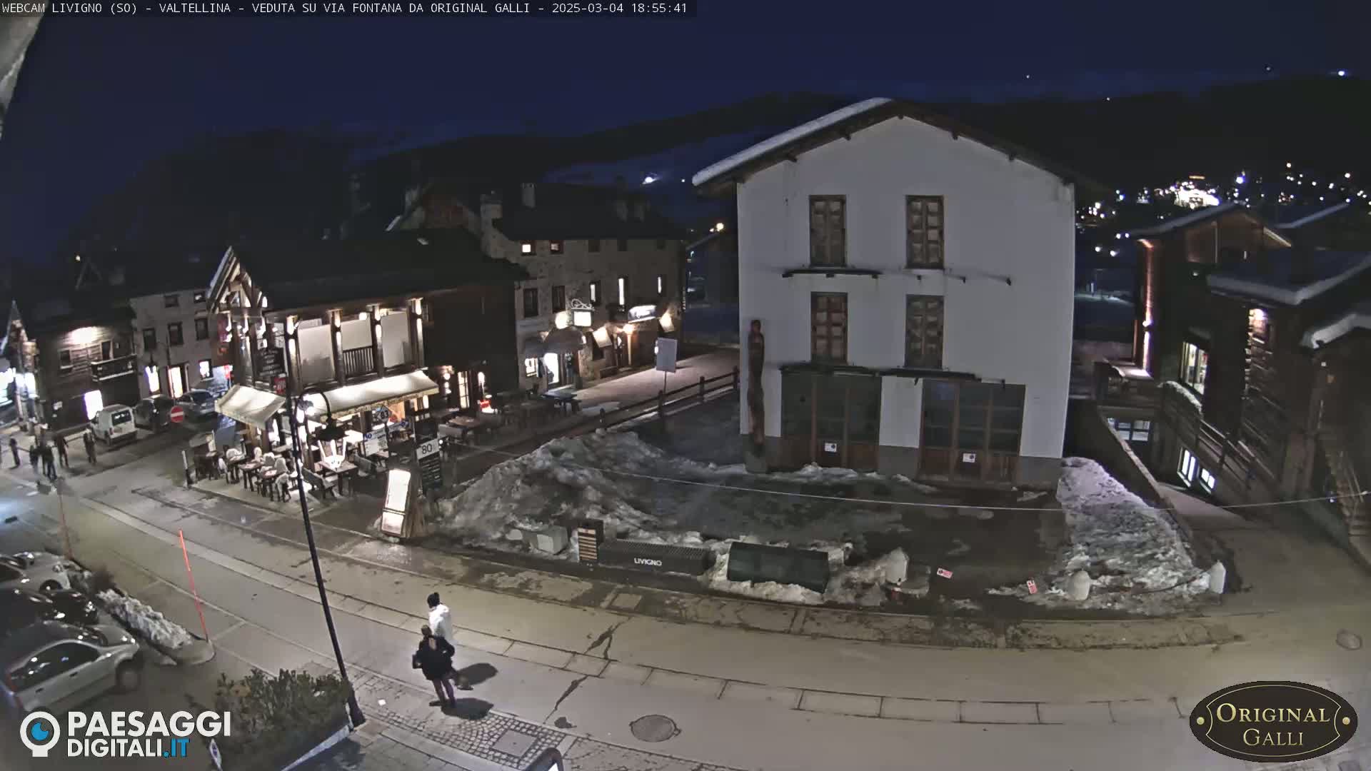 A nighttime street scene in a snow-dusted village shows several buildings, a few people walking, and cars parked along the road under a dark sky.