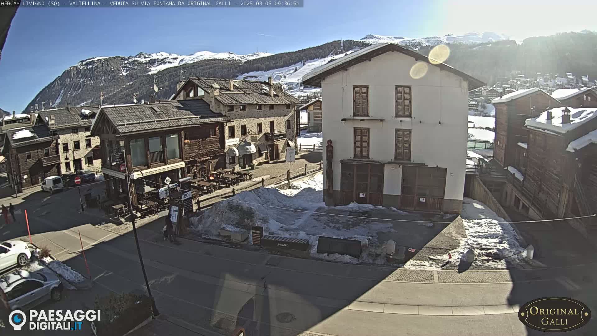 A sunny, snow-dusted village square is viewed from above, showing several buildings, a few cars, and patches of snow on the ground.