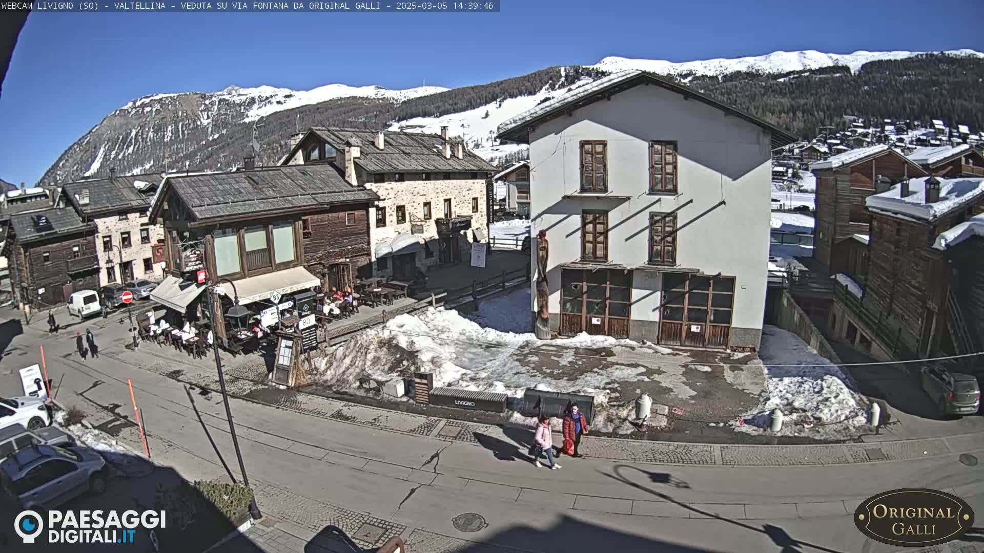 A sunny day in a snow-dusted village square, with several buildings, parked cars, and pedestrians.