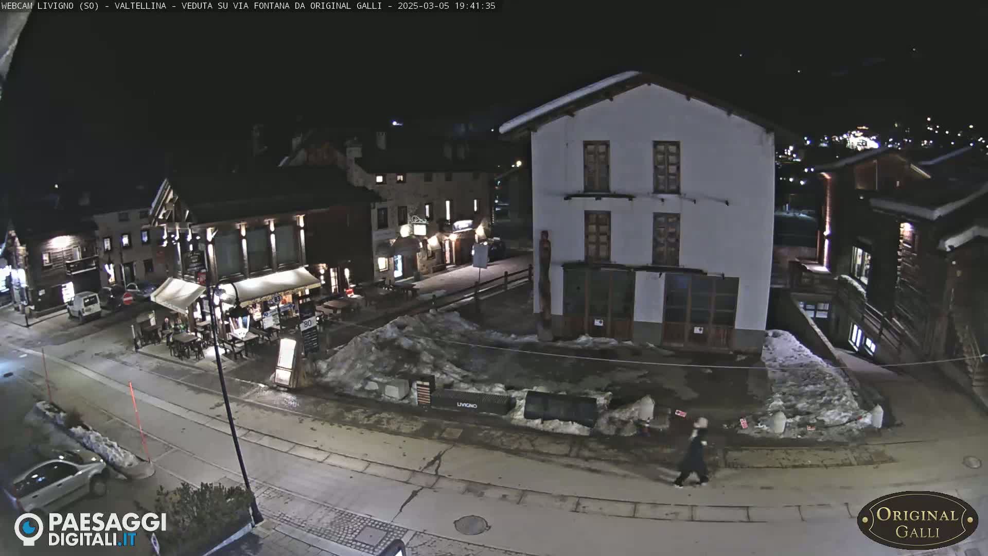 A nighttime view of a snow-dusted village street scene with several buildings, one of which is a two-story structure, and a person walking.